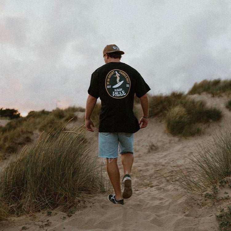 Man walking on a sandy path through grassy dunes wearing a black t-shirt with a logo.