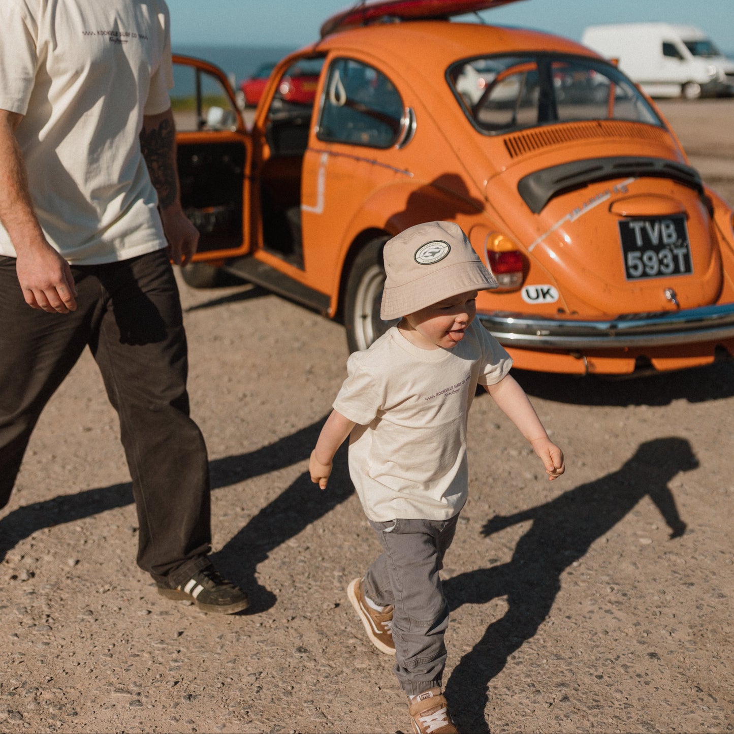 Man and child standing next to an orange vintage car with a surfboard on a beach.