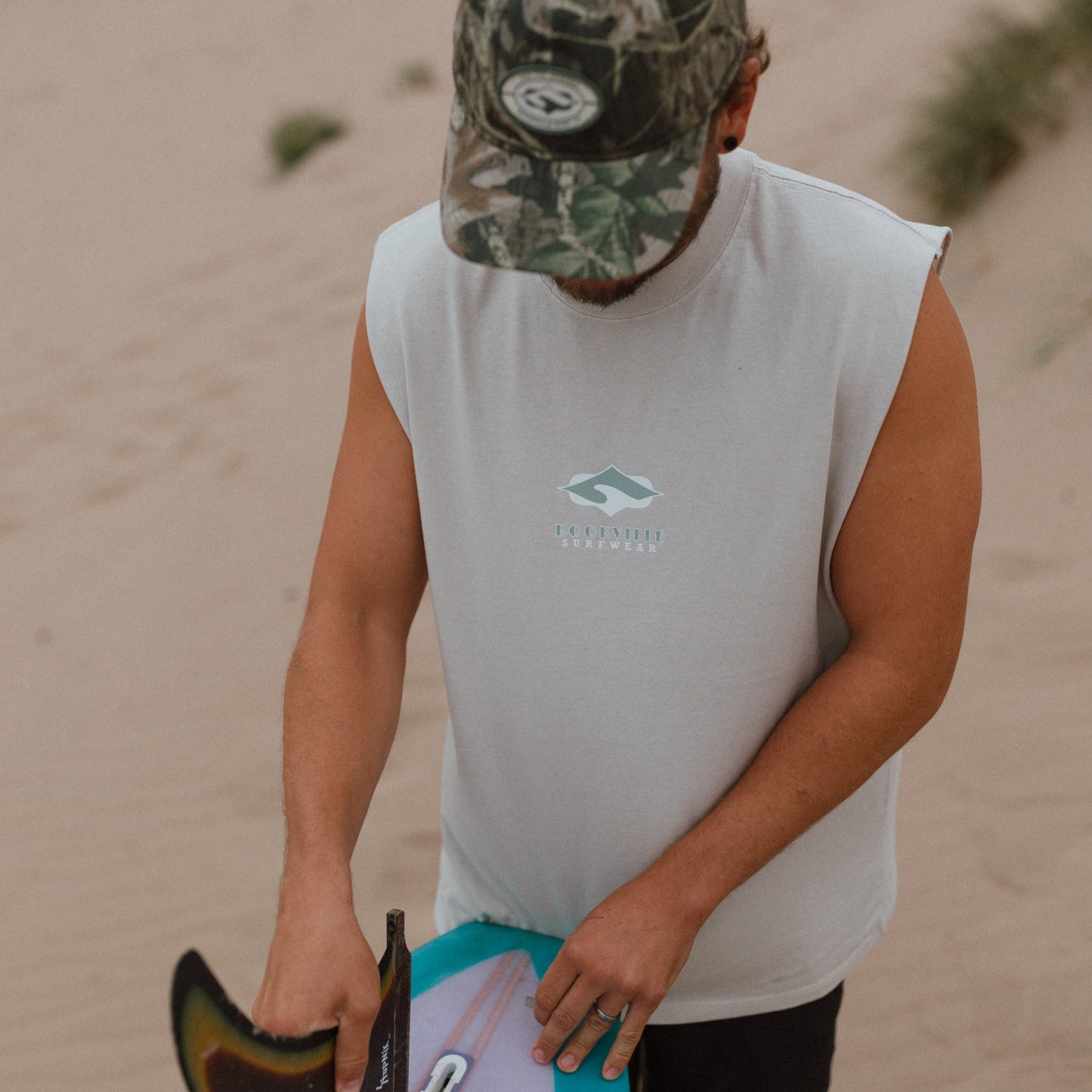 A man on a sandy beach wearing a white sleeveless shirt with a green mountain logo and a camouflage cap, holding a surfboard with a turquoise and white underside.