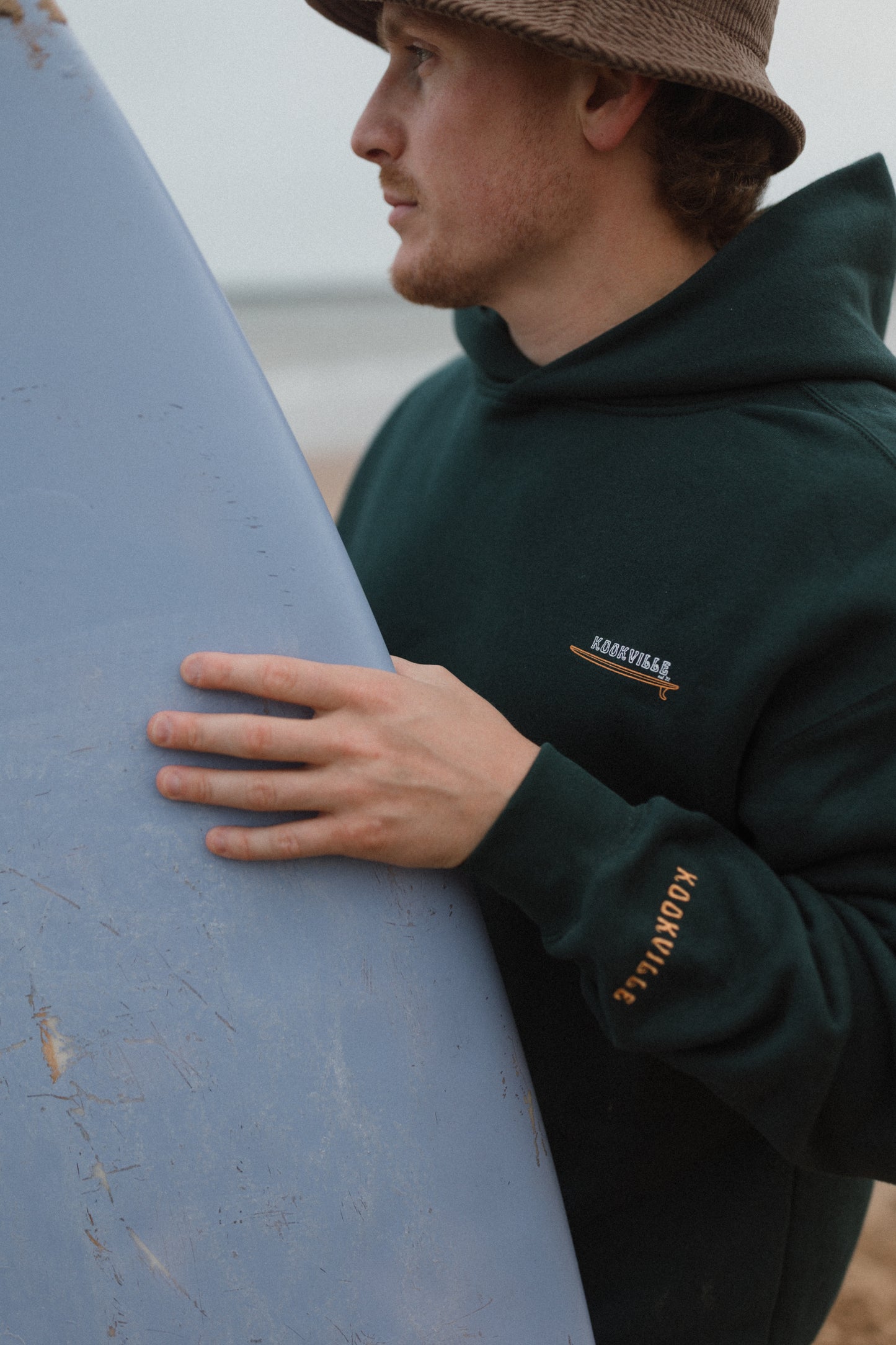 Man holding a surfboard wearing a green hoodie with branding on a beach.