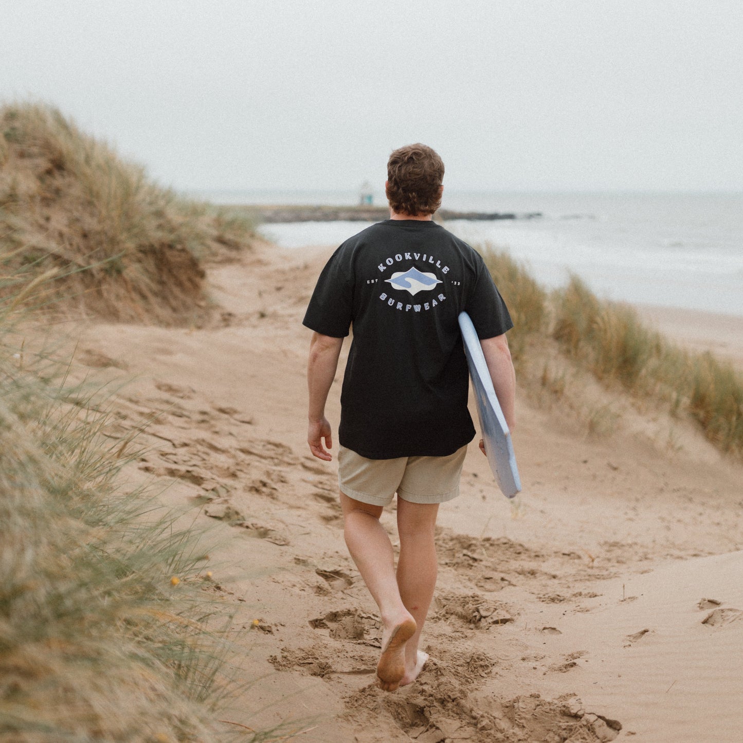 Man walking on a beach with a surfboard, wearing a black t-shirt with a logo.