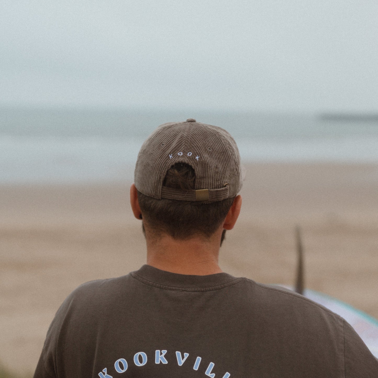 Person wearing a brown 'kookville Surfwear' t-shirt on a beach