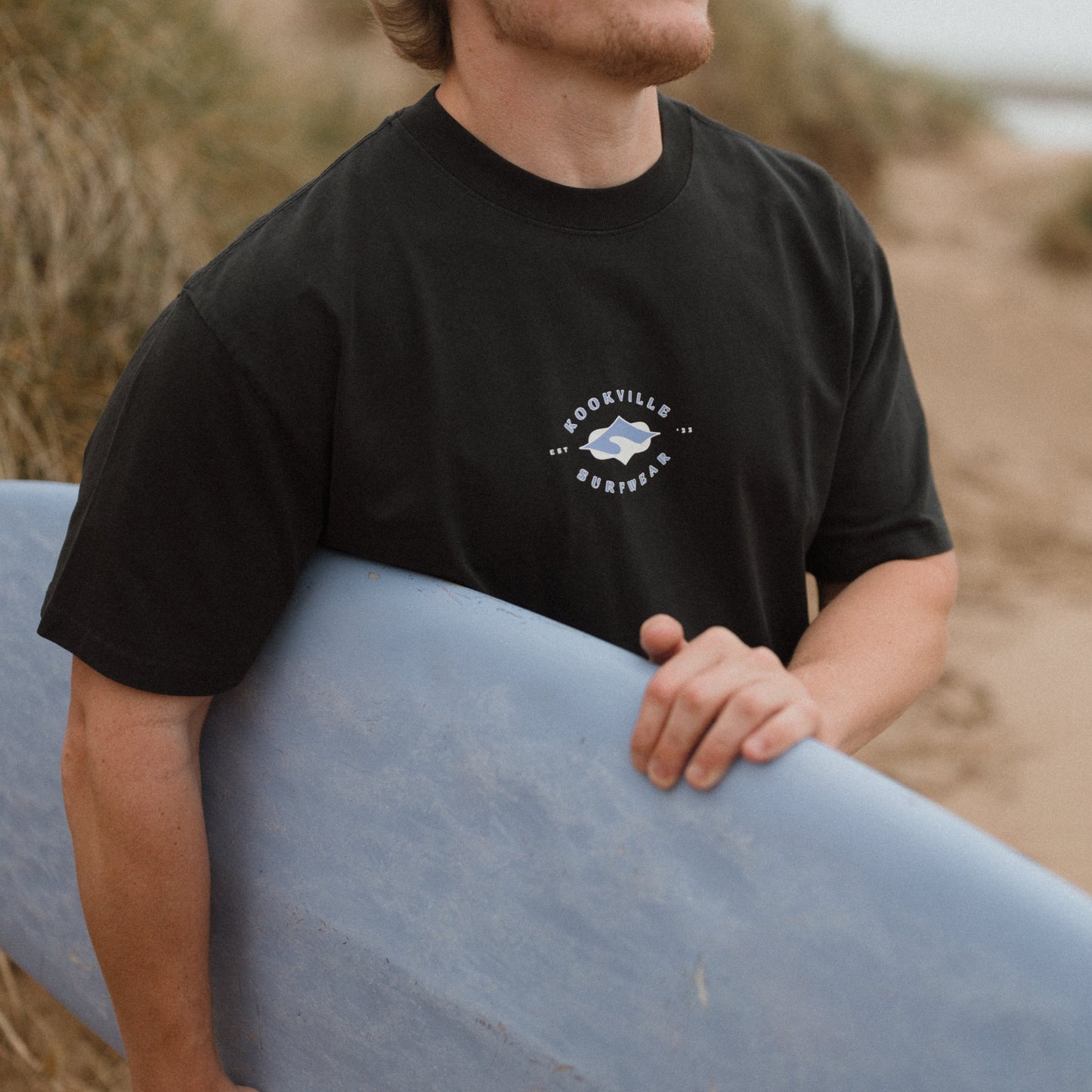 Person holding a surfboard on a beach with a black t-shirt featuring kookville surewear logo in purple and yellow