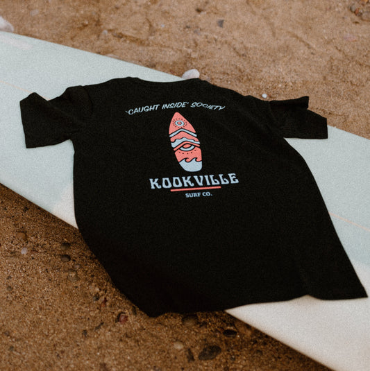 A black short-sleeved t-shirt with a graphic print of a surfboard and the text 'CAUGHT INSIDE SOCIETY' on the front, laid flat on a sandy surface with rocks in the background.