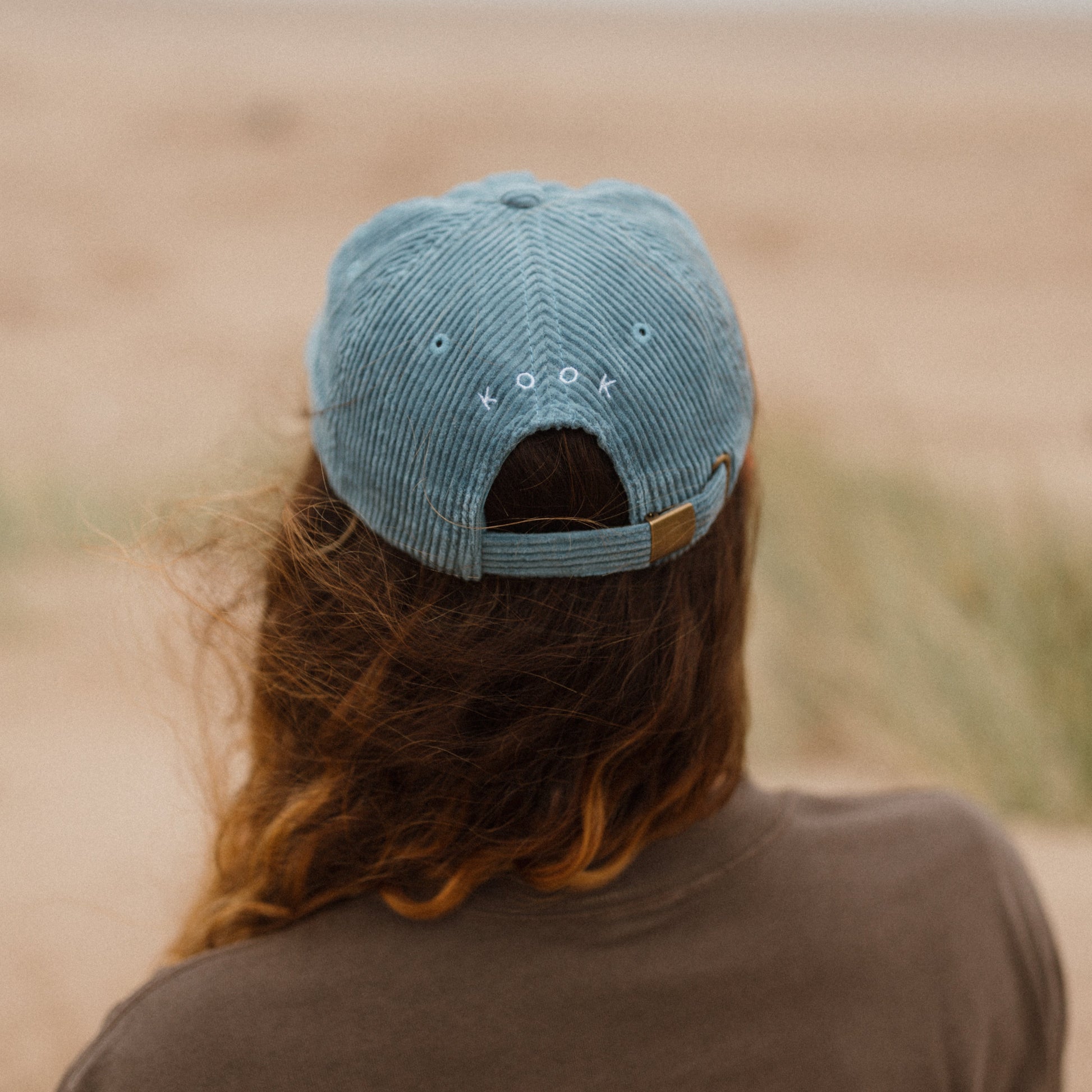 Woman wearing a blue cap and brown t-shirt with 'Kookville' text, standing on the beach