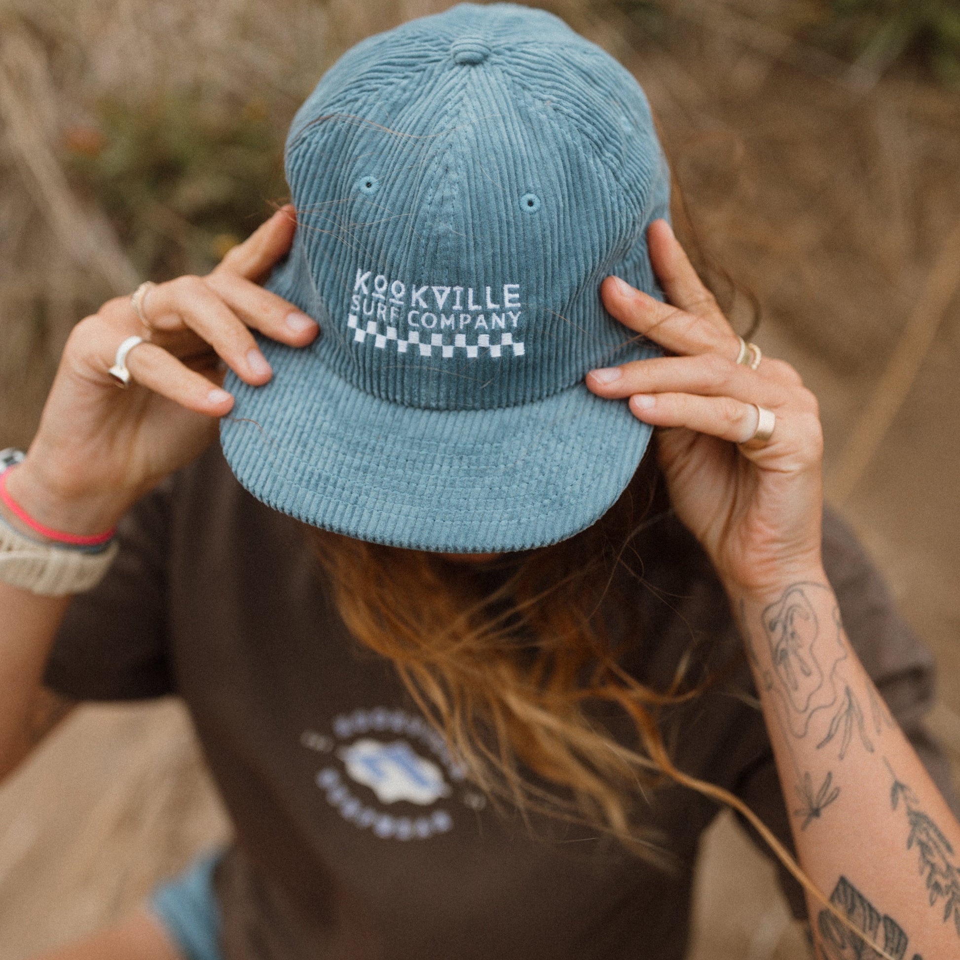 Person wearing a blue cap with 'Kookville' text, sitting on the beach.