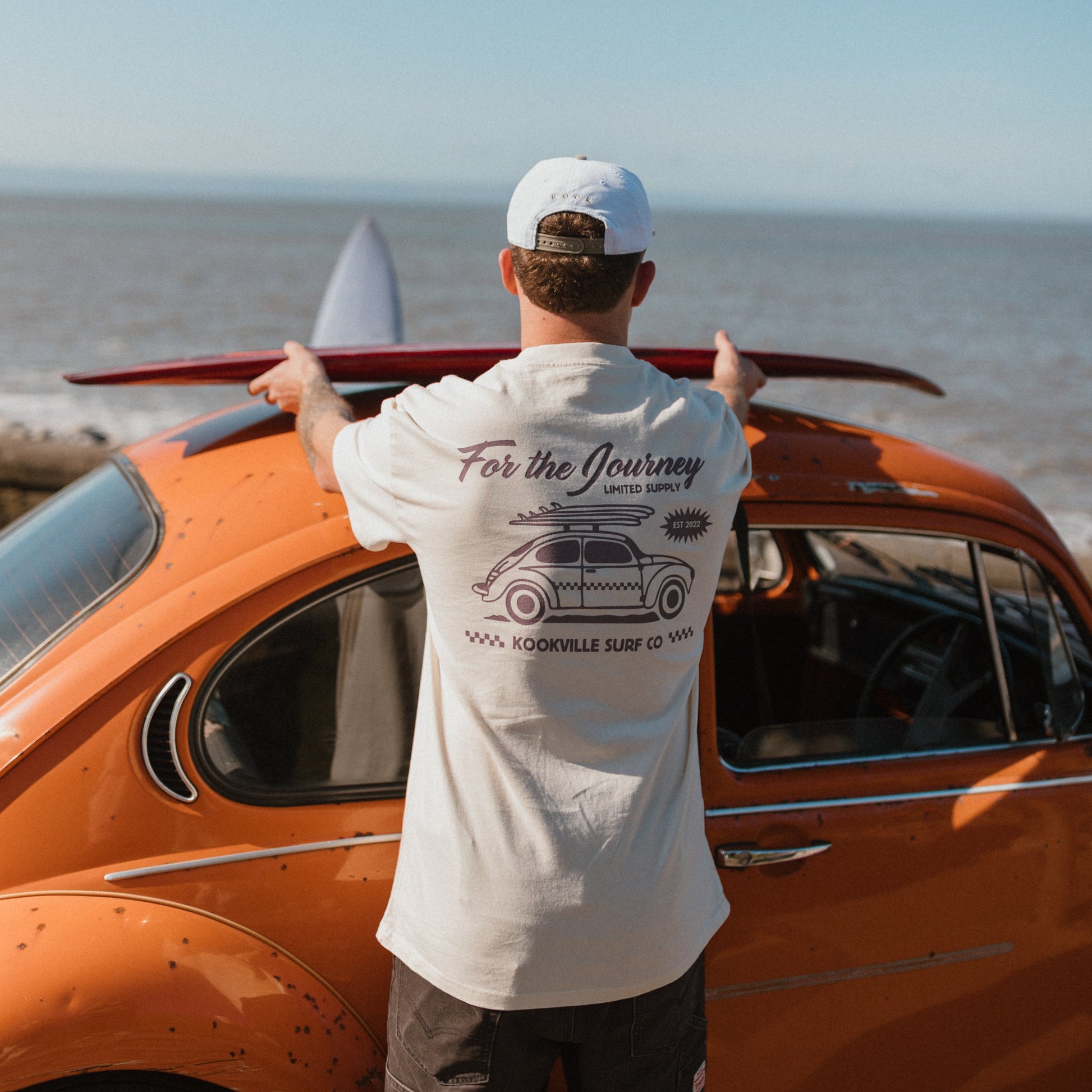 Man with a surfboard standing next to an orange car by the sea.