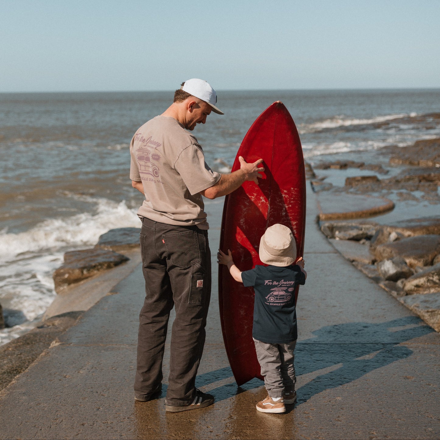 Man and child with a red surfboard on a rocky beach