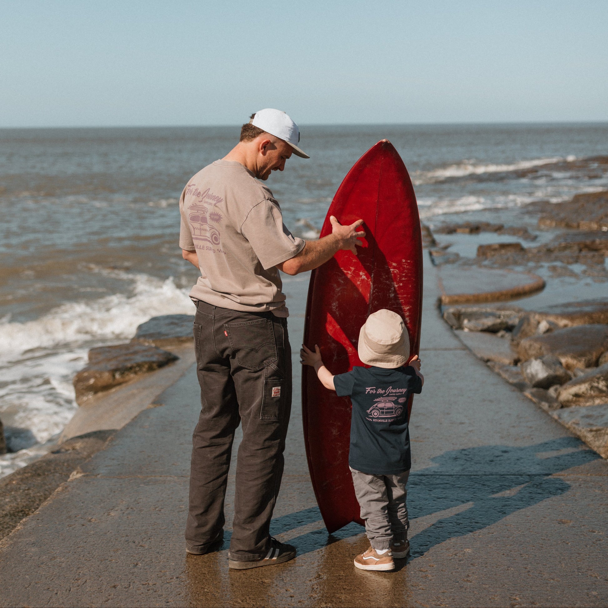 Man and child with a red surfboard on a rocky beach