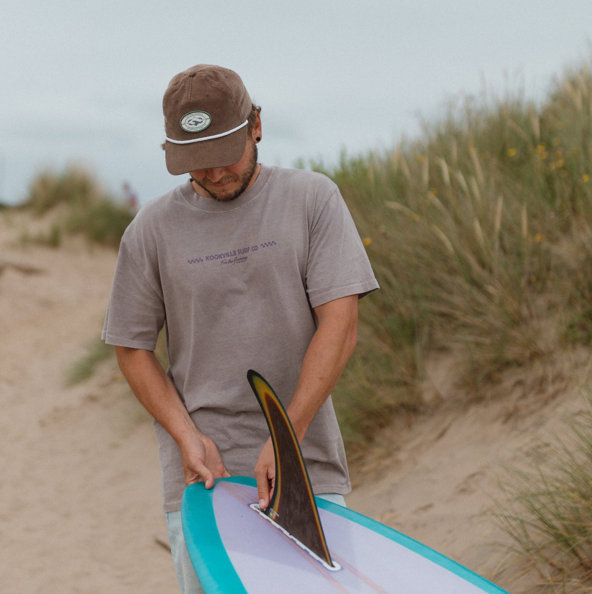 Man holding a surfboard on a sandy beach with grass and sky in the background