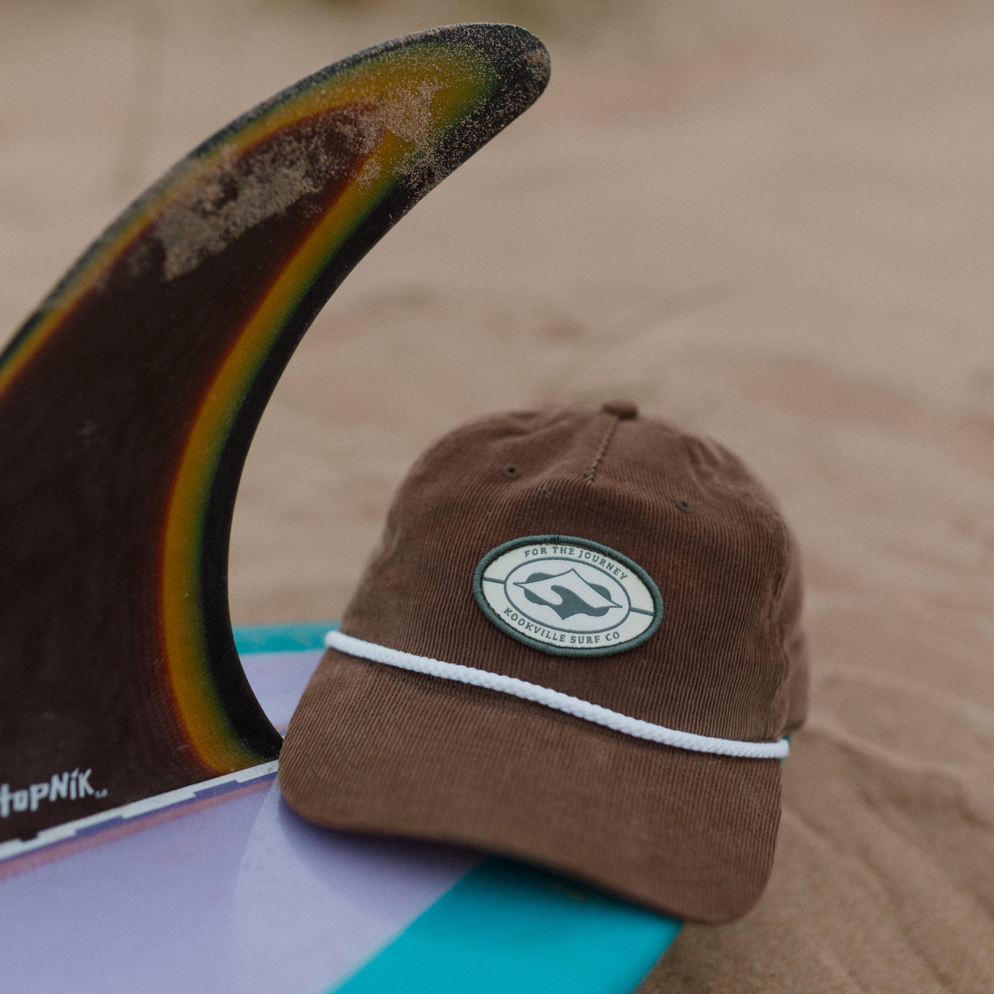 Brown cap with a logo on a surfboard fin against a sandy background