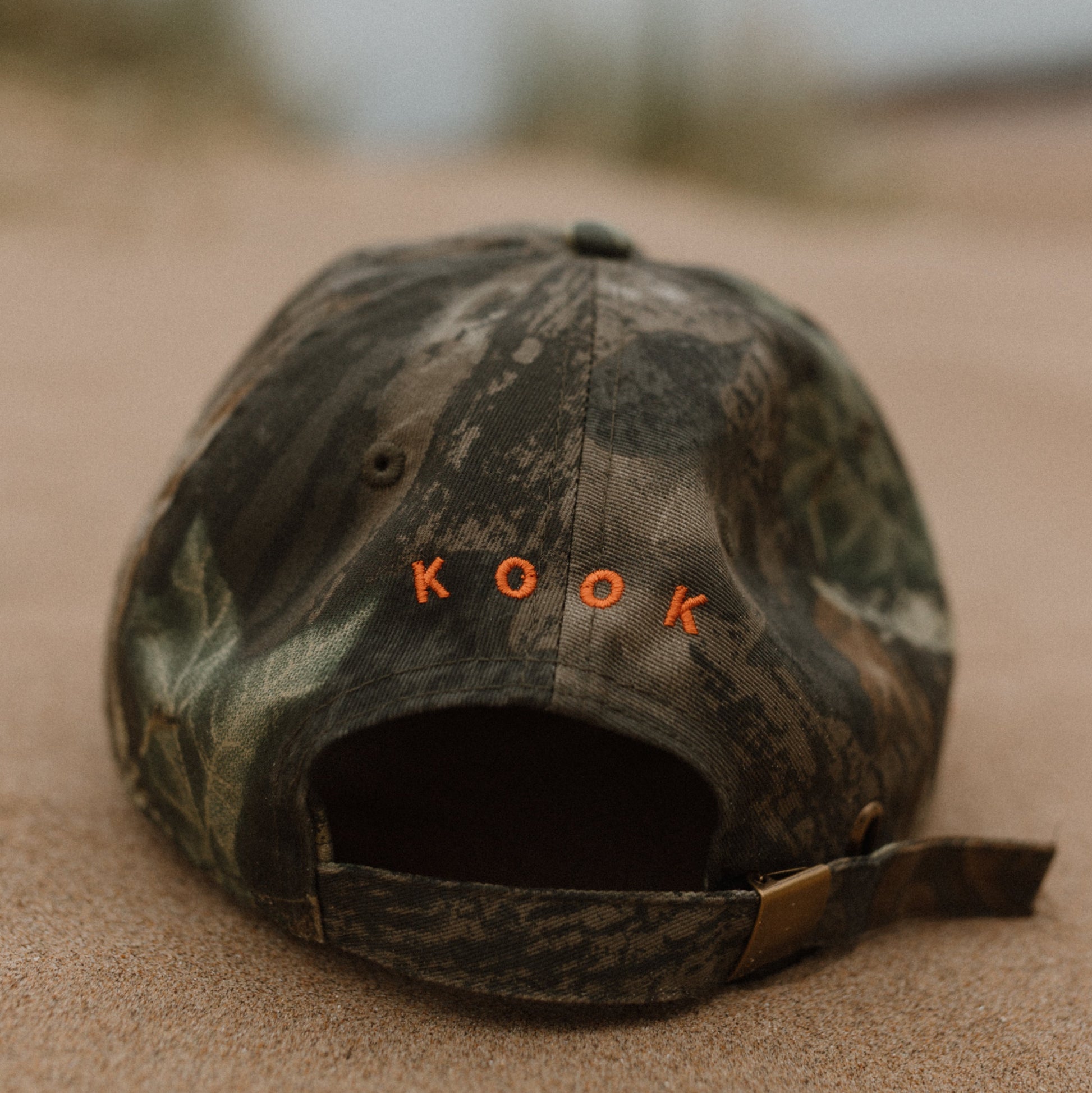 A woodland baseball cap with the word 'KOOK' embroidered in orange across the front, resting on sand with a blurred outdoor background.