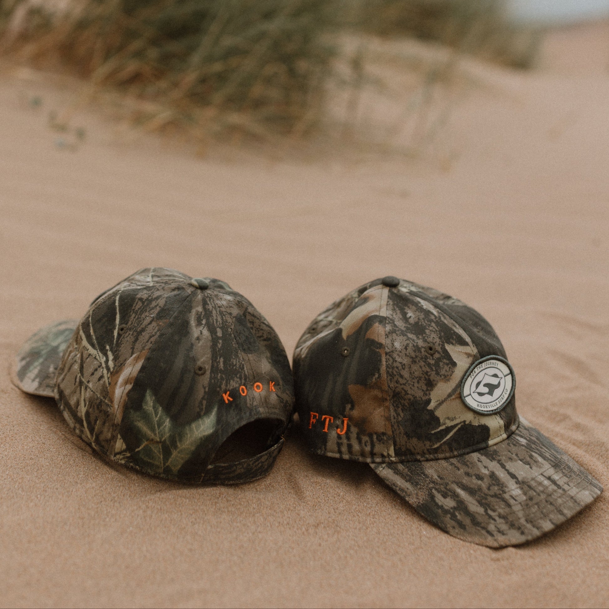Two camouflage baseball caps resting on sandy dunes with beach grass blurred in the background. One cap has orange embroidered text and a circular white patch on the front, and the other is shown from the side.