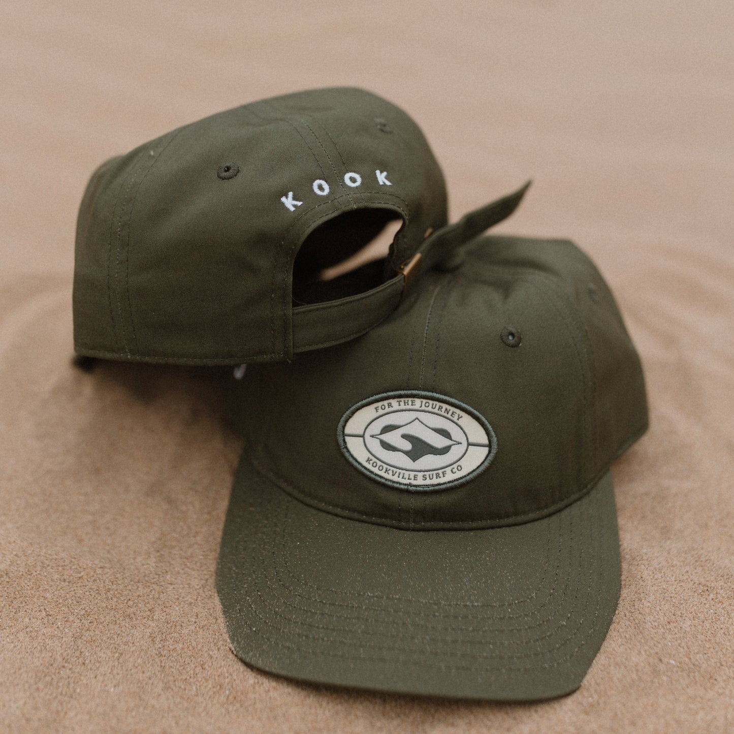 Two olive green baseball caps resting on sand, with one cap showing a curved brim and a circular white logo patch on the front, and the other cap angled behind it with the word 'KOOK' embroidered on the side.