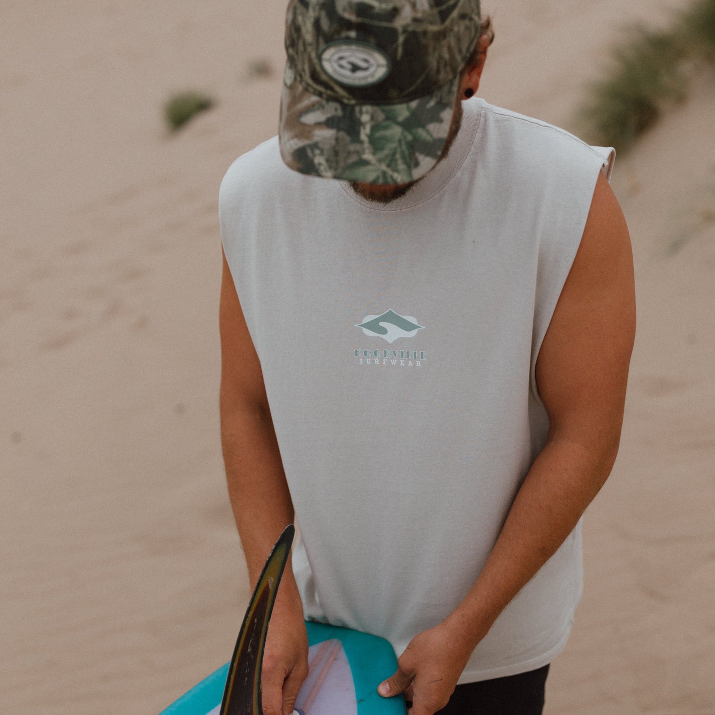 A man standing in sand wearing a camouflage baseball cap, a white sleeveless T-shirt with a small teal logo, and black shorts, holding a turquoise and white surfboard.