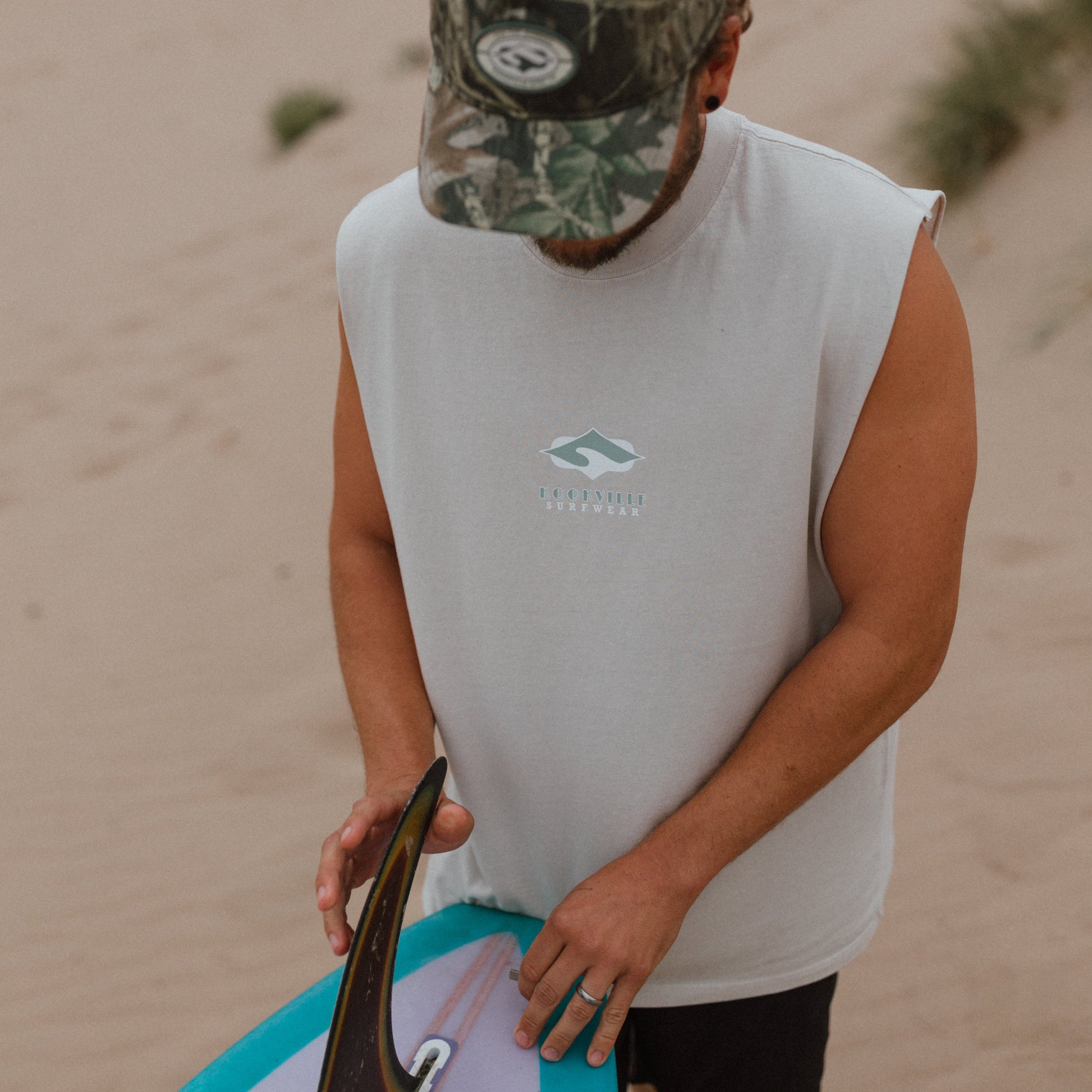 A man on a sandy beach wearing a white sleeveless surf shirt with a small blue and white logo on the chest, a camouflage baseball cap, and black shorts, holding a surfboard with a turquoise and white underside.
