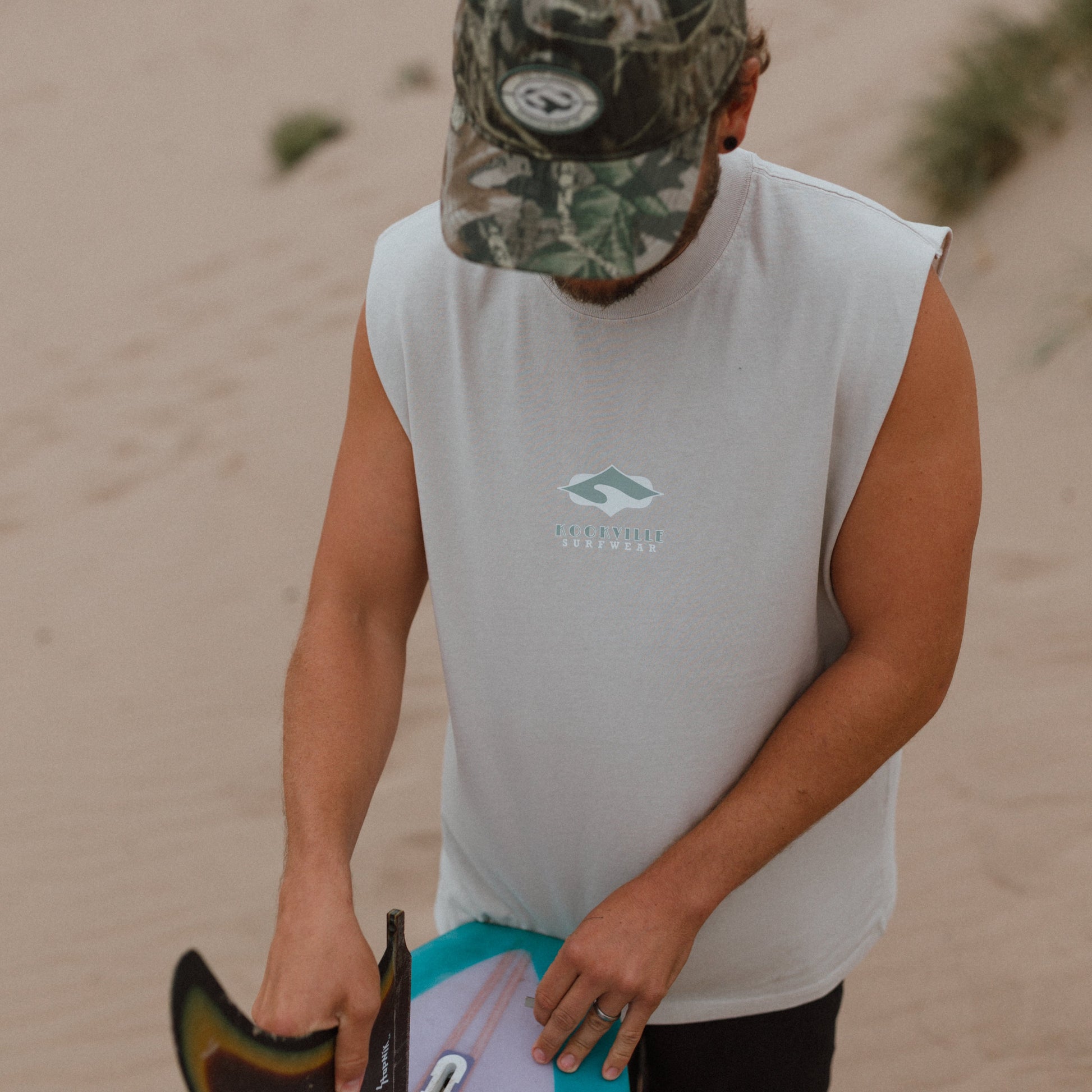A man on a sandy beach wearing a white sleeveless shirt with a green mountain logo and a camouflage cap, holding a surfboard with a turquoise and white underside.
