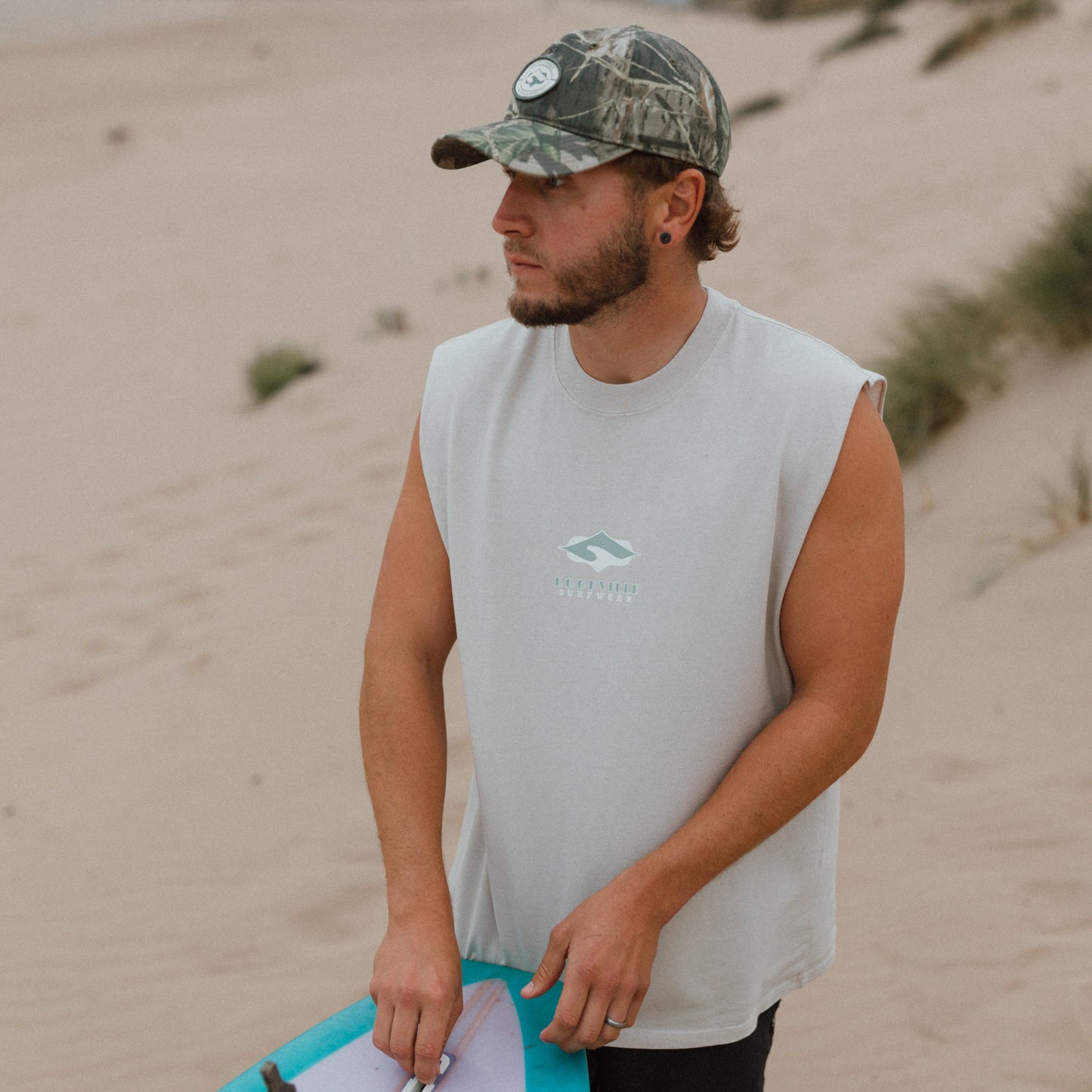 A man standing on a sandy beach wearing a white sleeveless t-shirt, black shorts, and a camouflage baseball cap, holding a pink and turquoise surfboard.