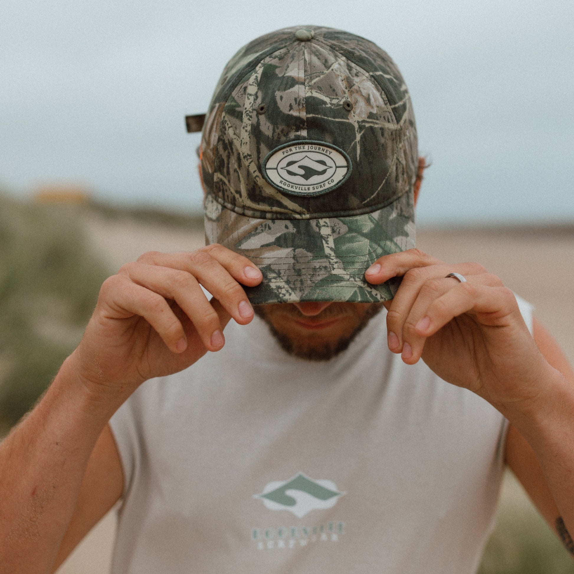 A man on a beach wearing a Realtree camouflage baseball cap and a light gray sleeveless shirt with a green and white logo. He is holding the brim of the cap down over his eyes.
