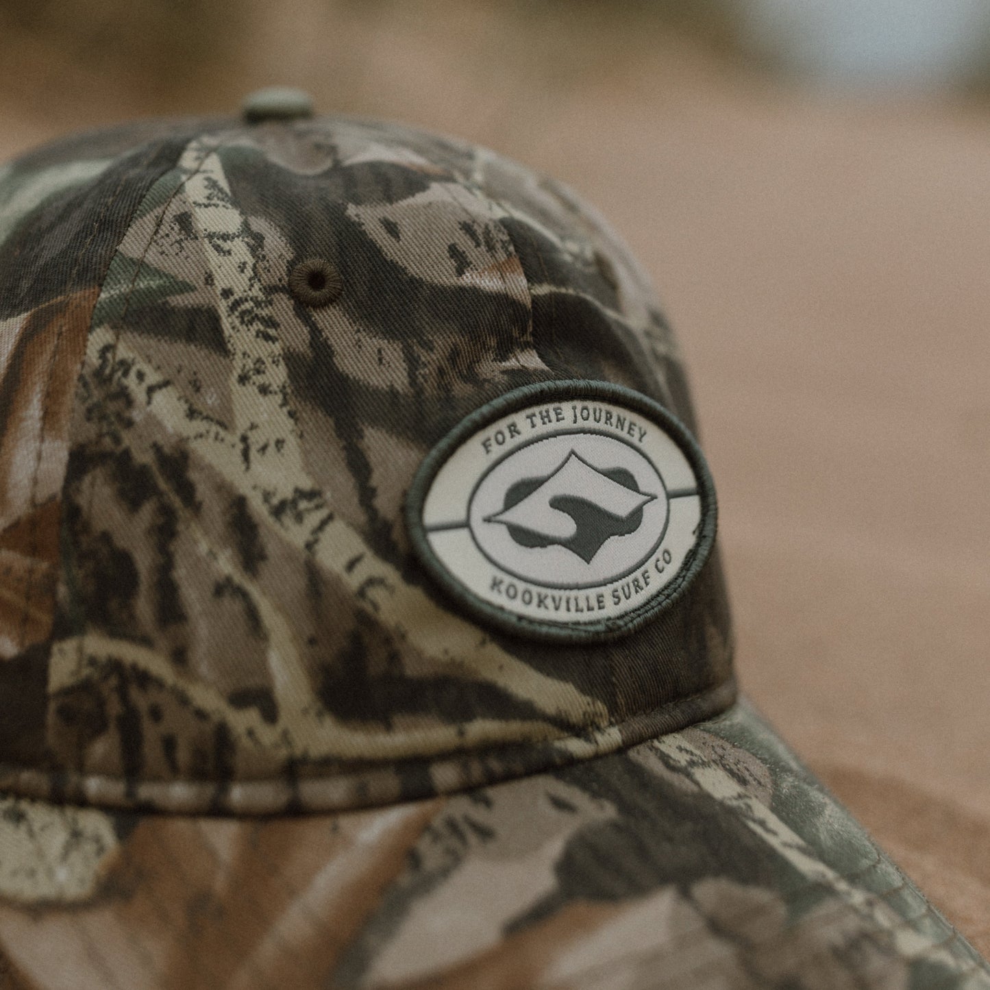 A close-up of a woodland baseball cap with a circular embroidered patch reading 'For The Journey' and 'Kookville Surf Co.' The background is a blurred outdoor sandy environment.