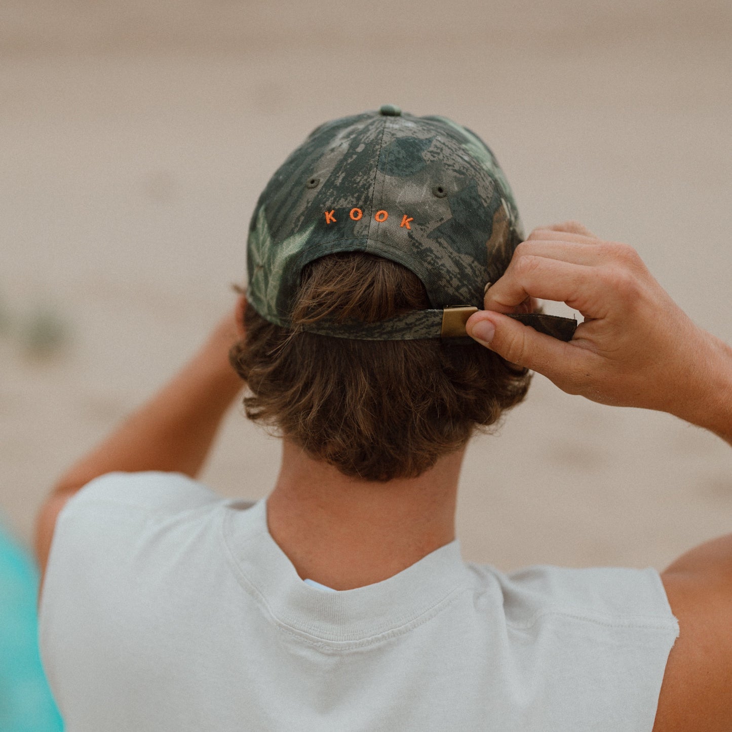 A man viewed from behind on a beach, wearing a camouflage baseball cap with 'KOOK' embroidered in orange on the back and adjusting the strap.