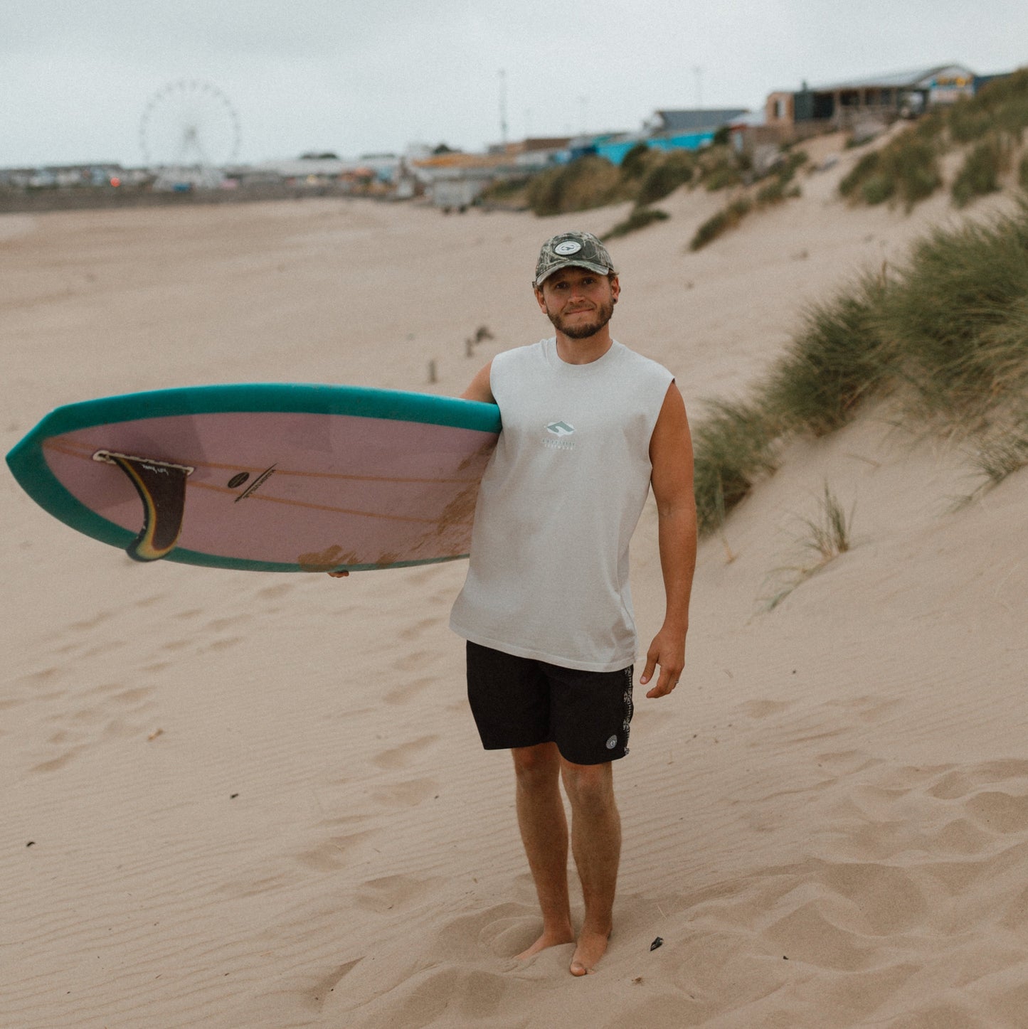 A man standing on a sandy beach holding a surfboard under his arm. He is wearing a white sleeveless shirt, black shorts, and a camouflage cap.