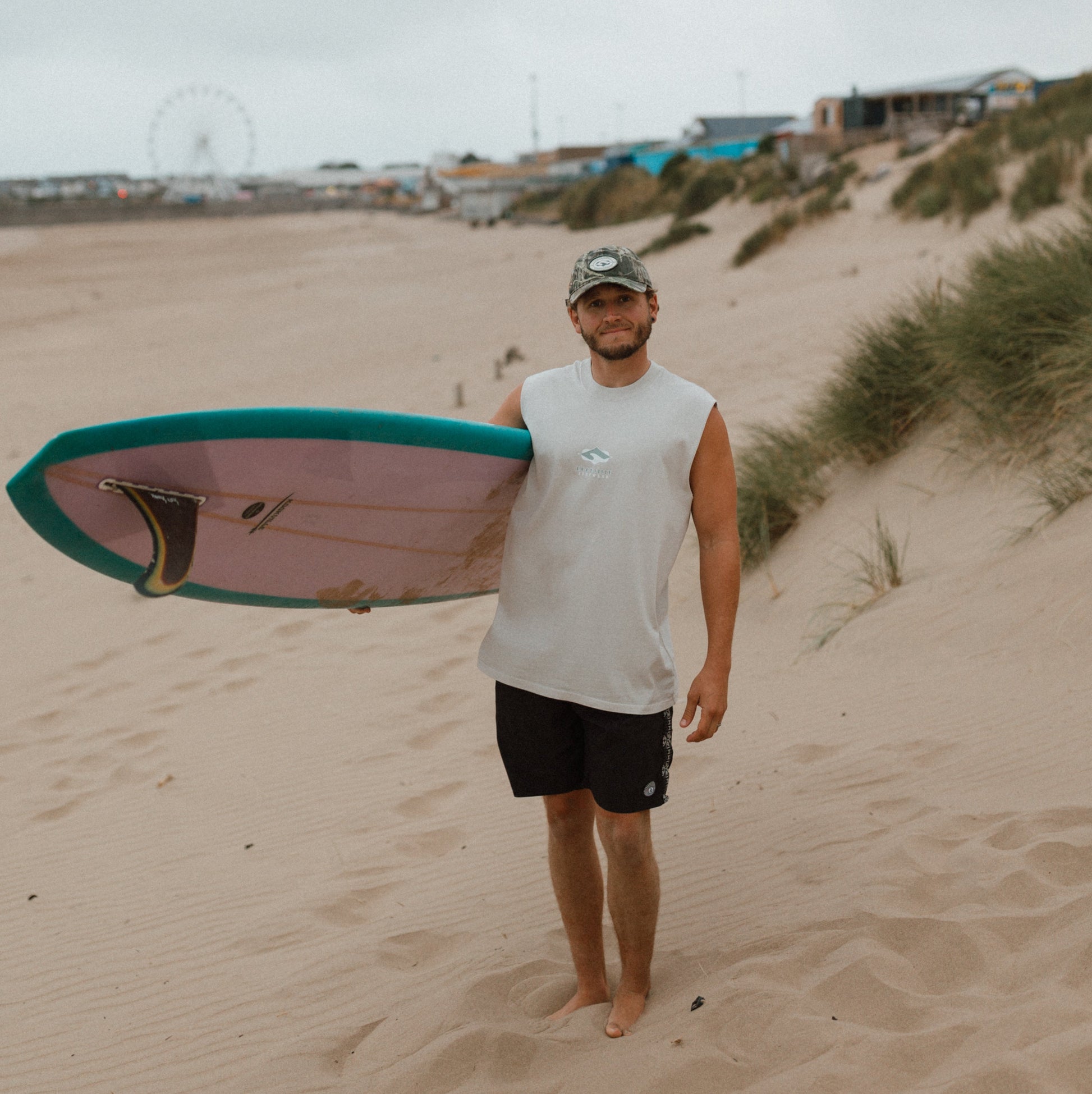 A man standing on a sandy beach holding a surfboard under his arm. He is wearing a white sleeveless shirt, black shorts, and a camouflage cap.