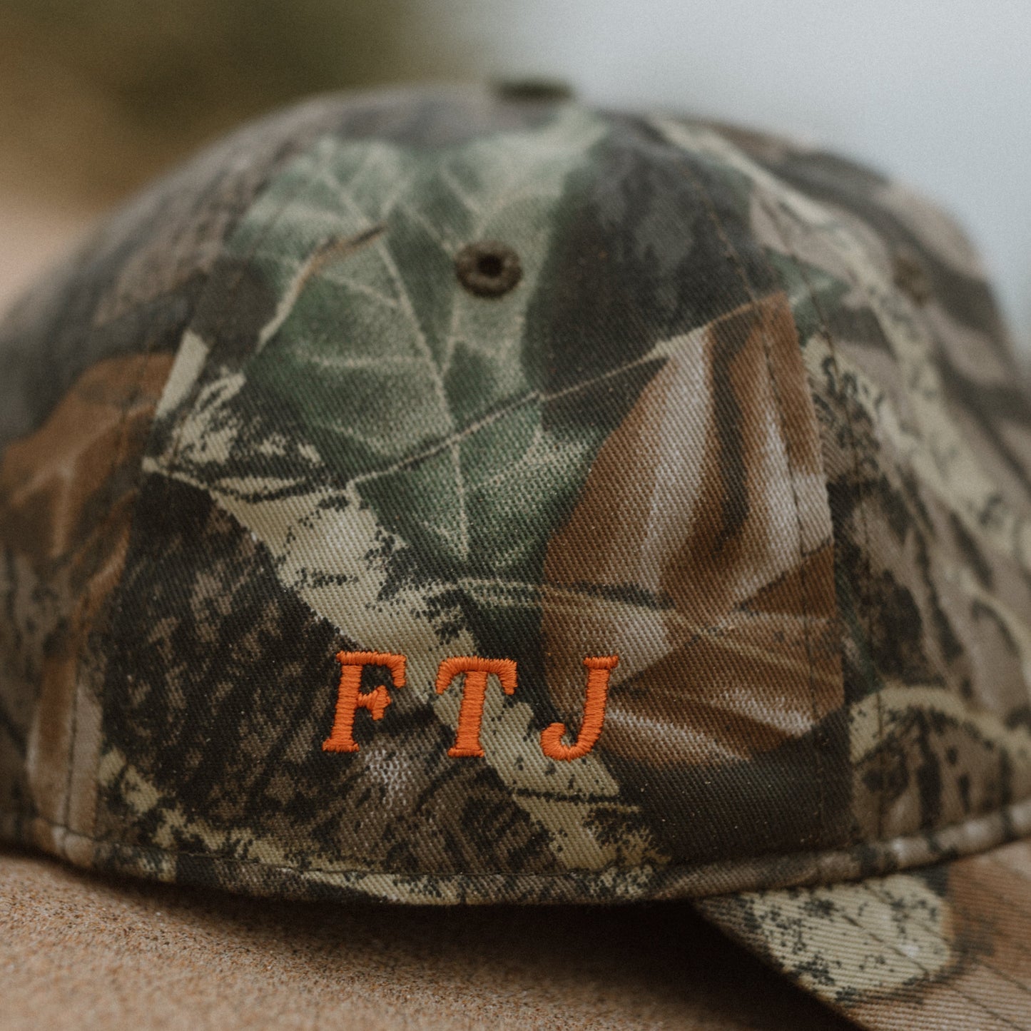 A close-up of a camouflage baseball cap with orange embroidered letters 'FTJ' on the front, resting on a sandy surface with a blurred outdoor background.