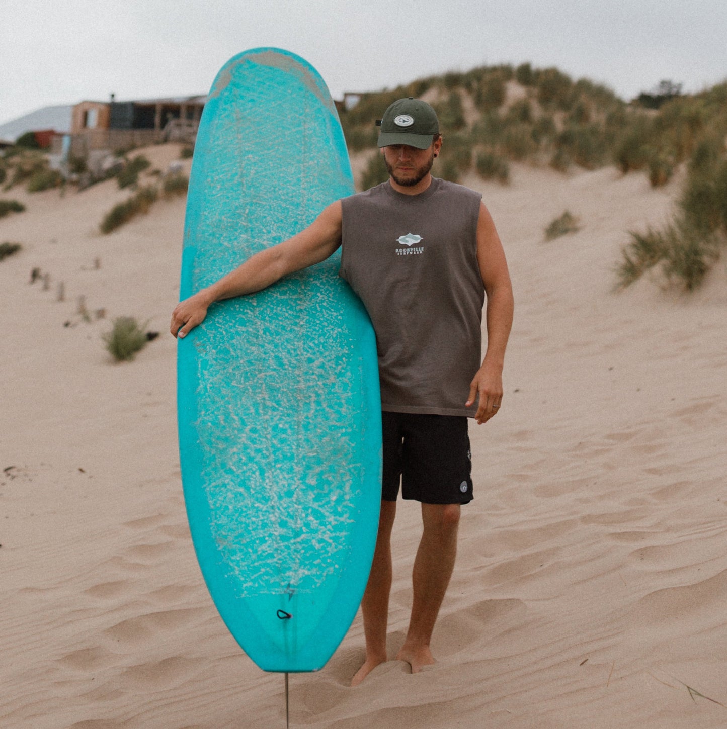 A man walking on sandy dunes carrying a bright turquoise surfboard under his arm, wearing a gray sleeveless shirt, black shorts, and a dark cap.