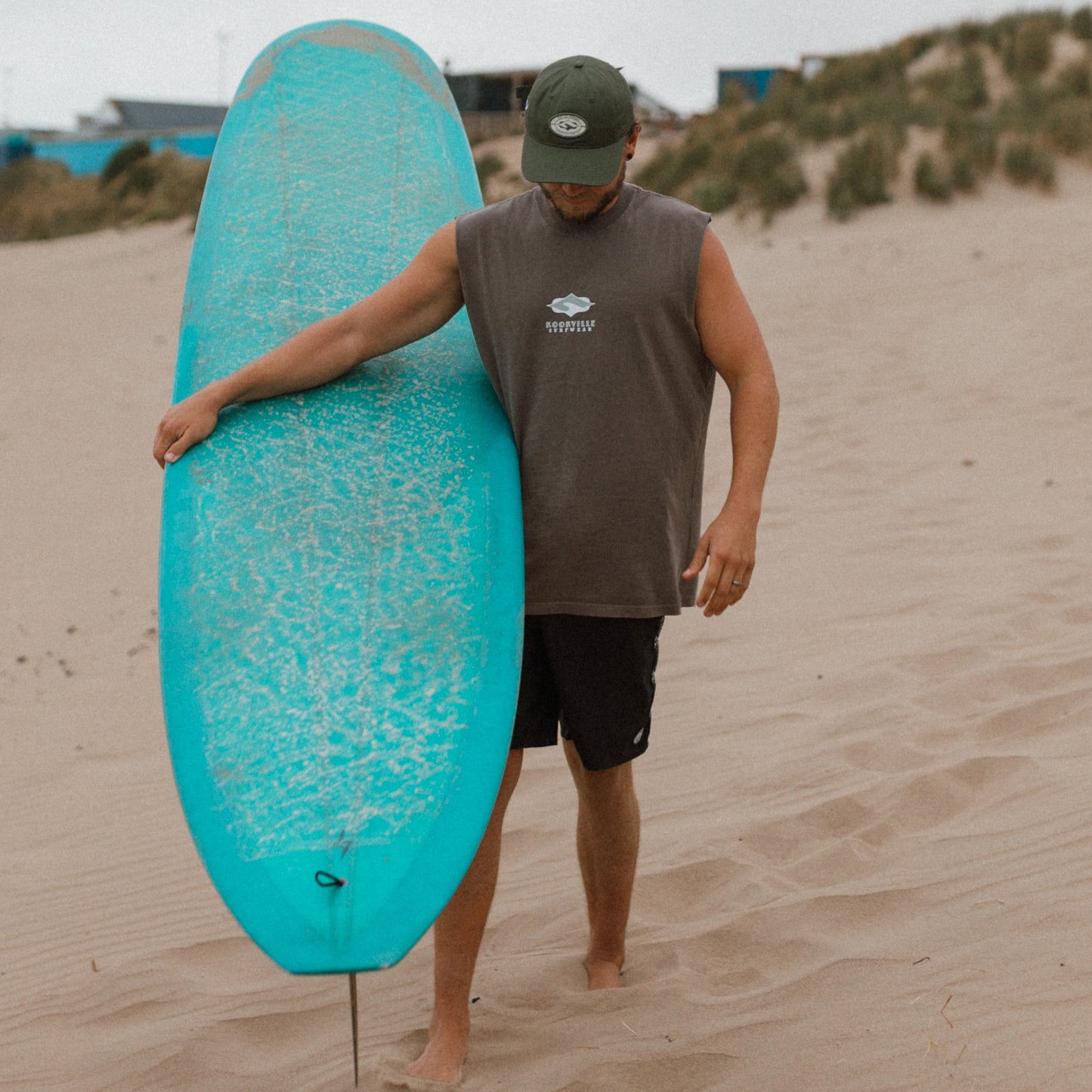 A man walking through sand dunes carrying a turquoise surfboard, wearing a brown sleeveless shirt, black shorts, and a dark green cap.