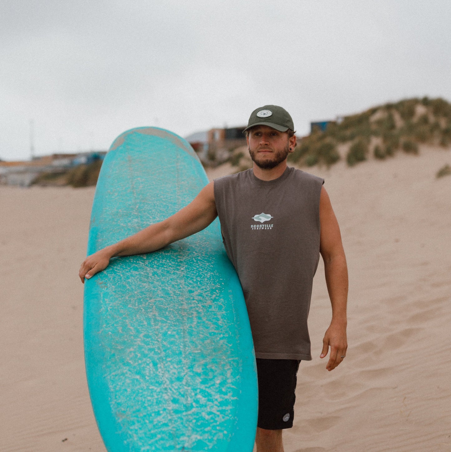 A man walking on a sandy beach carrying a bright turquoise surfboard under his arm, wearing a gray sleeveless shirt, black shorts, and a dark cap.
