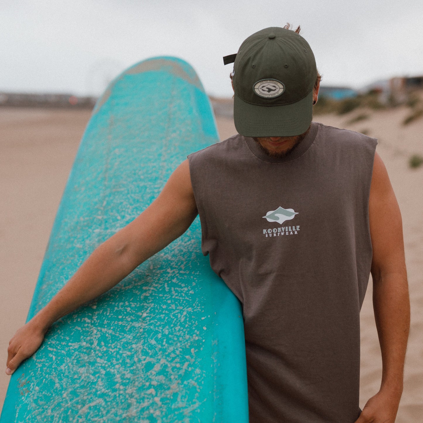 A man on a sandy beach carrying a turquoise surfboard under his arm, wearing a dark green cap, a sleeveless brown shirt with a white and green surf logo