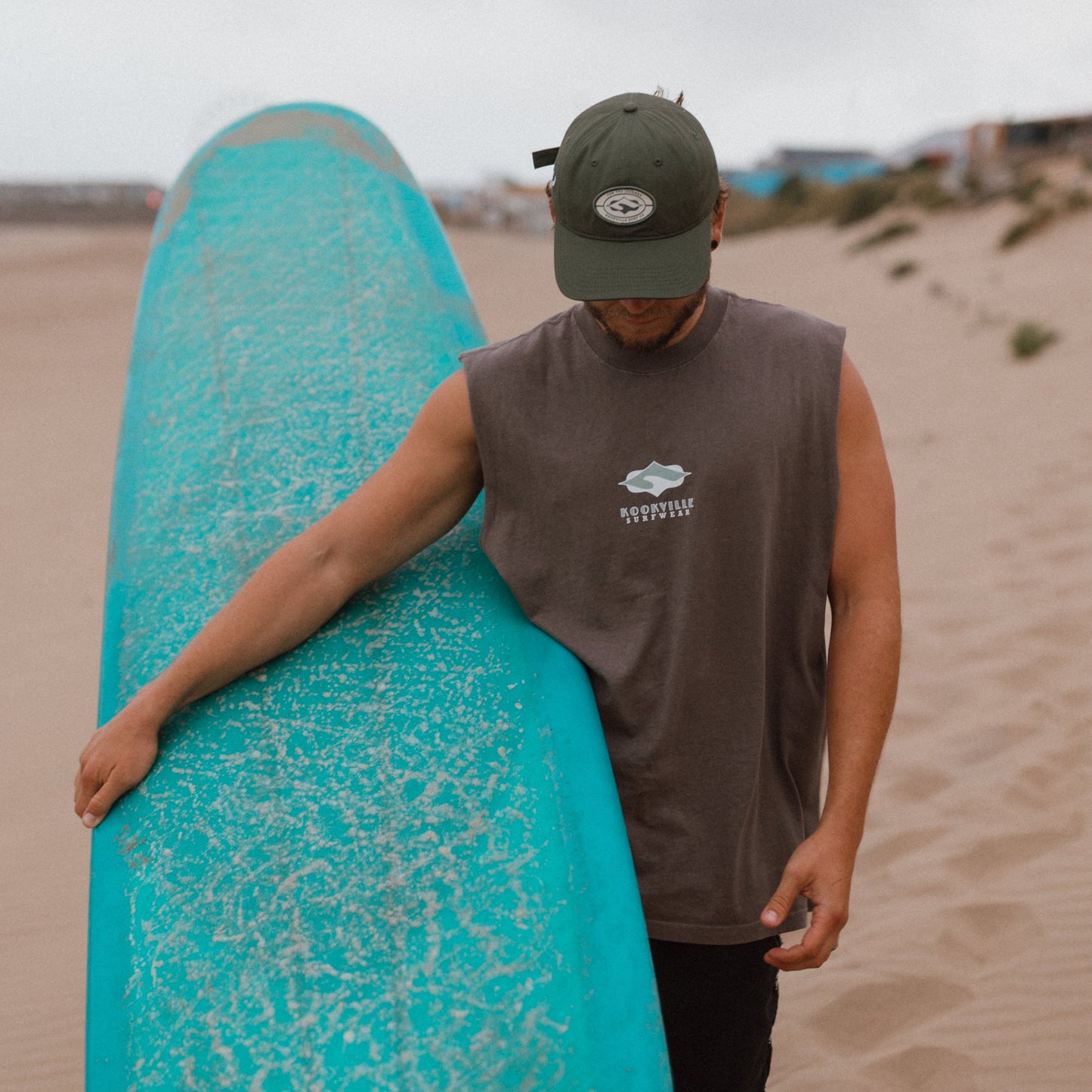 A man on a sandy beach carrying a large turquoise surfboard under his arm, wearing a dark green cap, a sleeveless grey t shirt with a white logo, and black shorts.