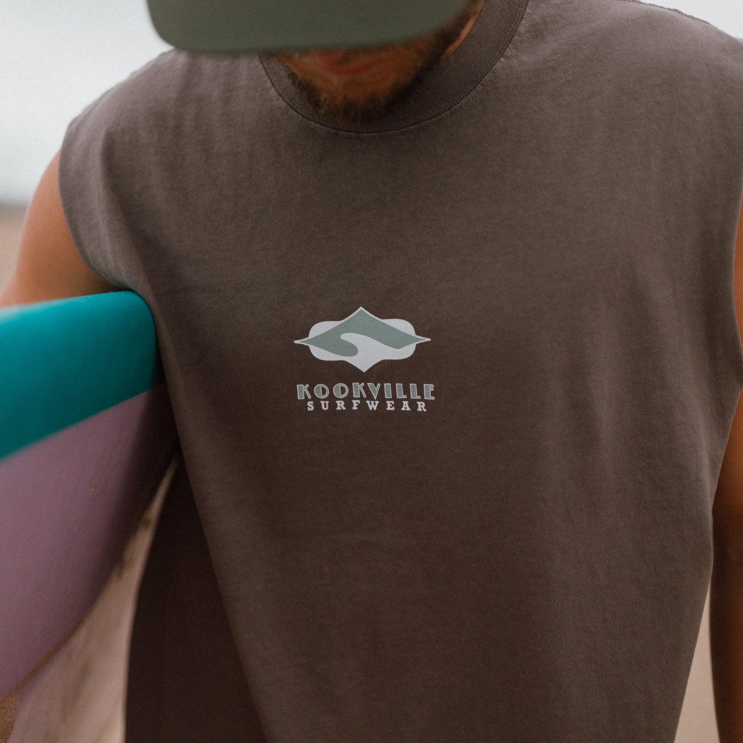 A close-up of a man wearing a sleeveless dark brown t-shirt with a white 'Kookville Surfwear' logo on the chest, holding a surfboard on a beach.