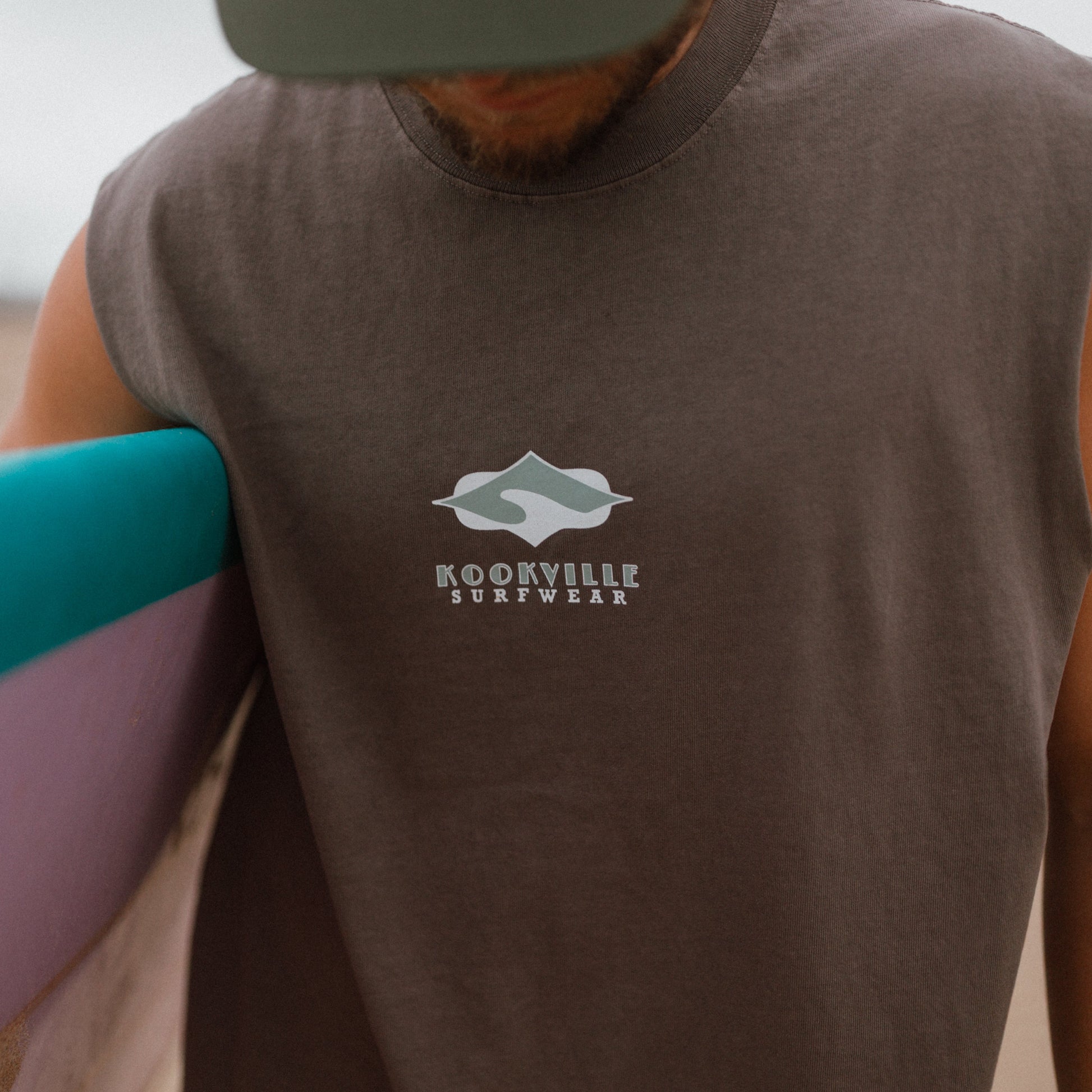 A close-up of a man wearing a sleeveless dark brown t-shirt with a white 'Kookville Surfwear' logo on the chest, holding a surfboard on a beach.