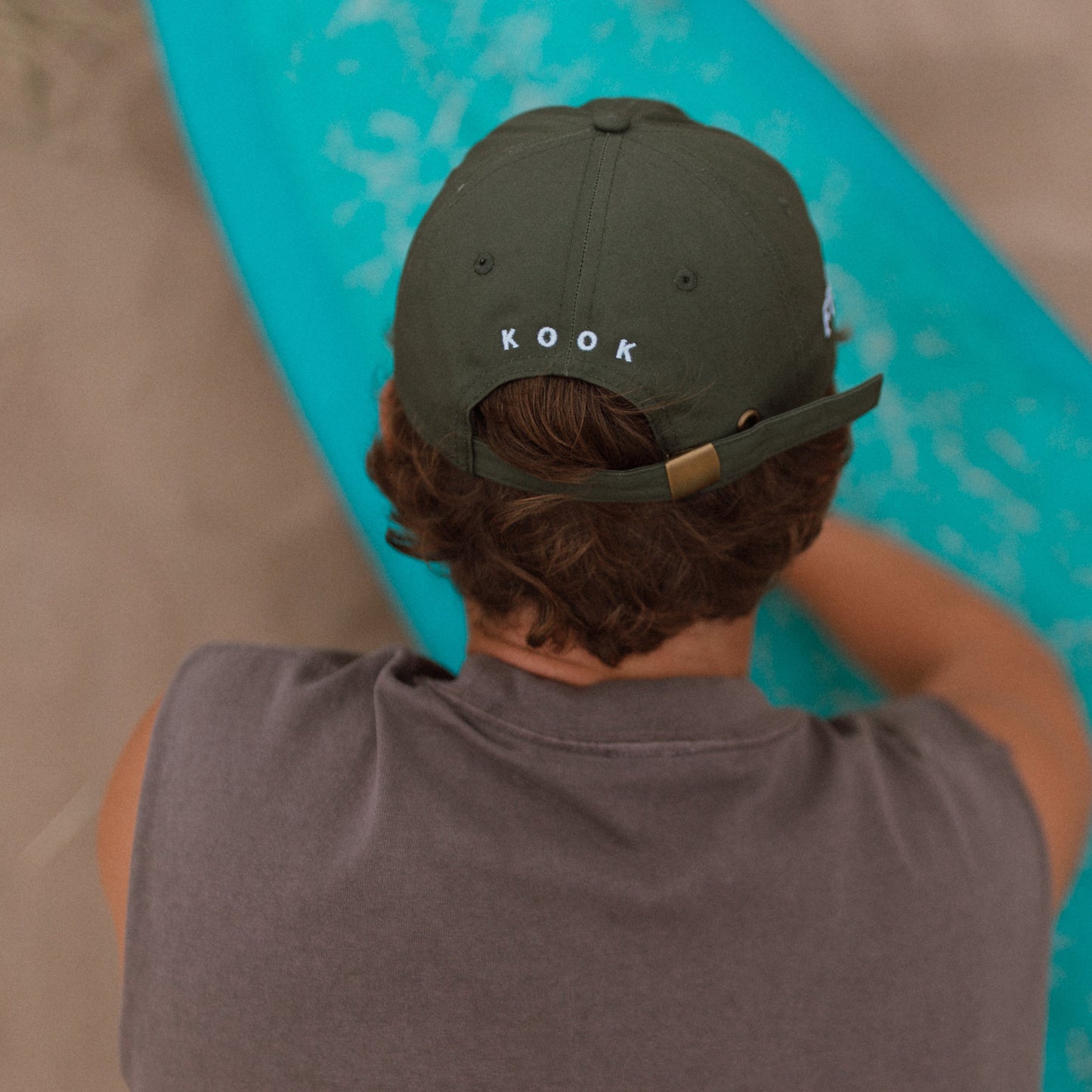 A person viewed from behind wearing an khaki green Kook brand baseball cap and a gray sleeveless shirt, holding a turquoise surfboard on a sandy beach.
