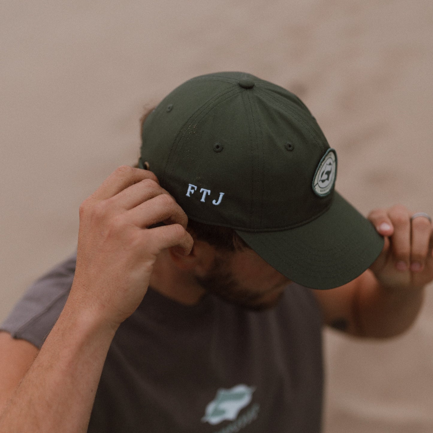 A man wearing an khaki green baseball cap with 'FTJ' embroidered on the side and a circular patch on the brim, paired with a dark gray t-shirt against a sandy beige background.