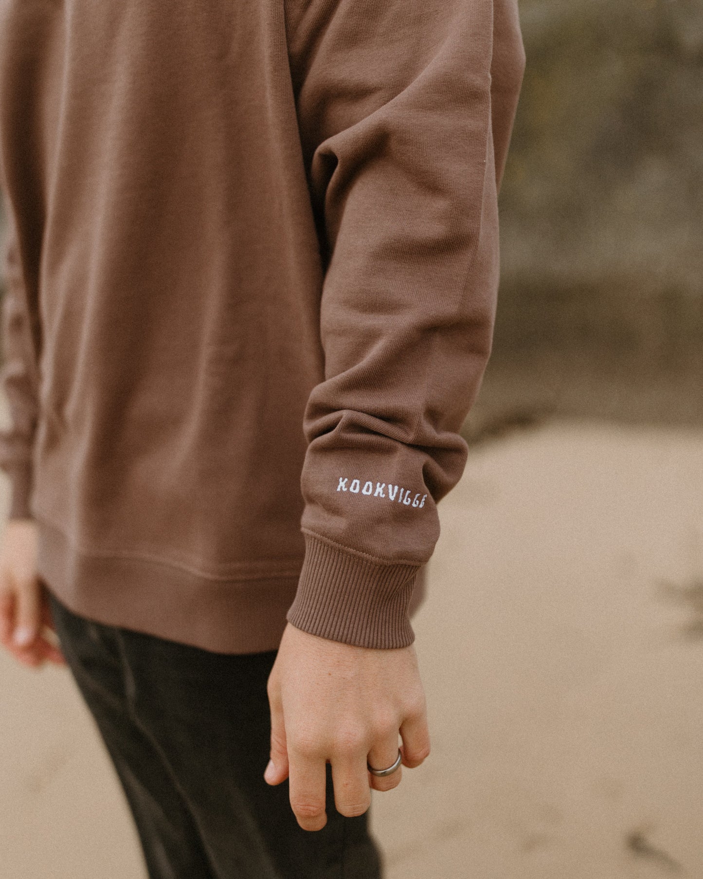 Brown sweatshirt with visible branding on sleeve against a blurred natural background