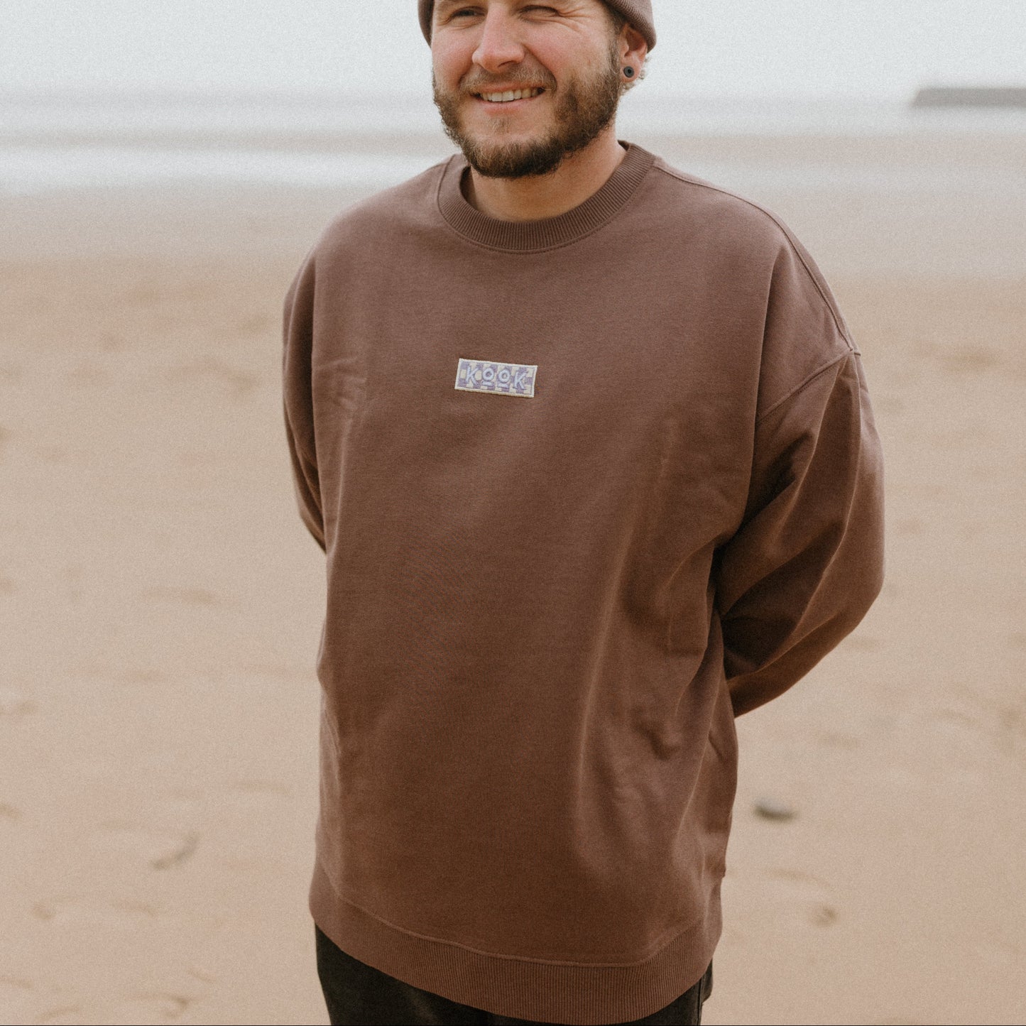 Man wearing a brown sweatshirt and beanie standing on a beach.
