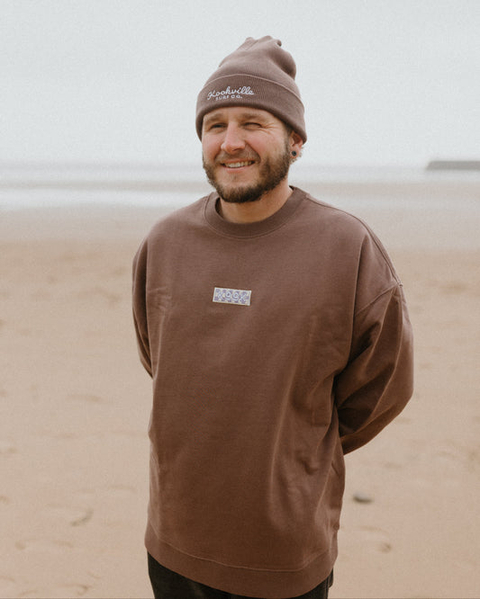 Man wearing a brown sweatshirt and beanie standing on a beach.