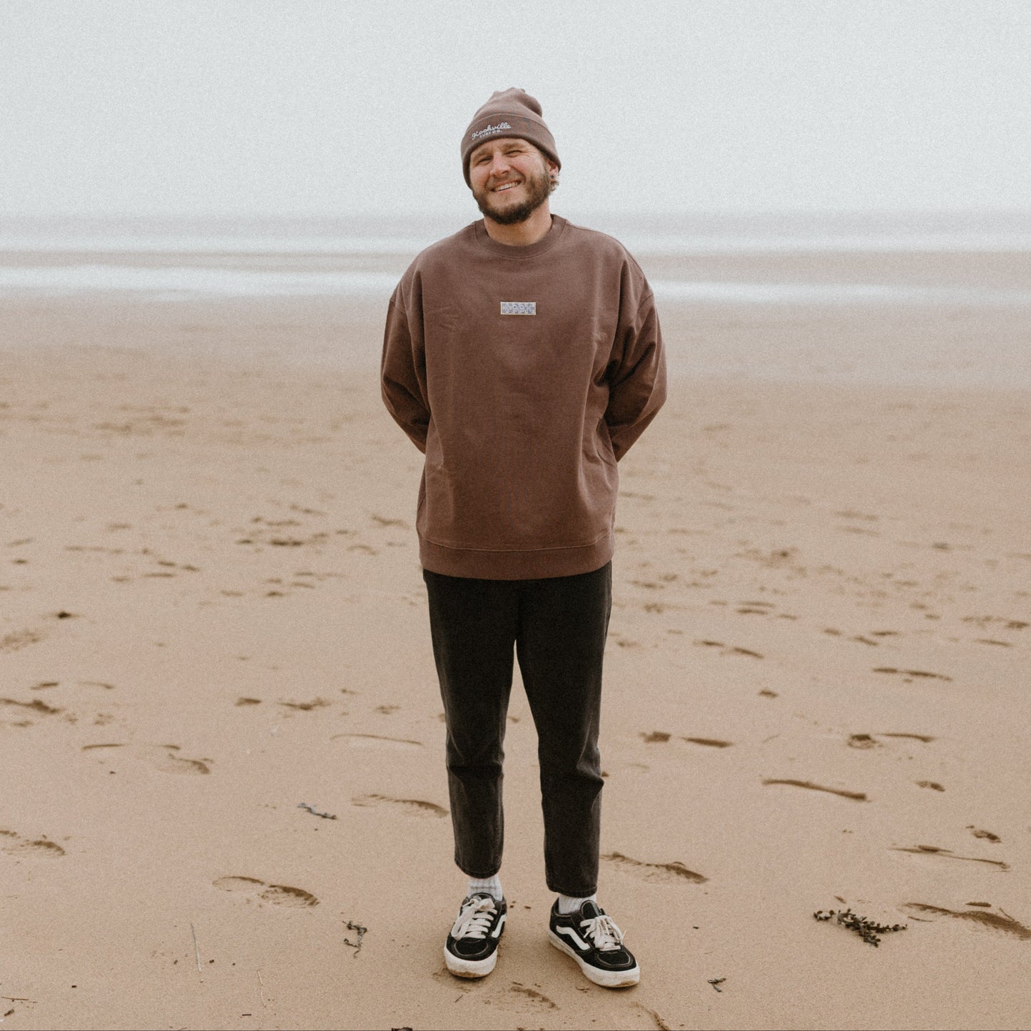 Man standing on a beach wearing a brown sweatshirt and beanie.