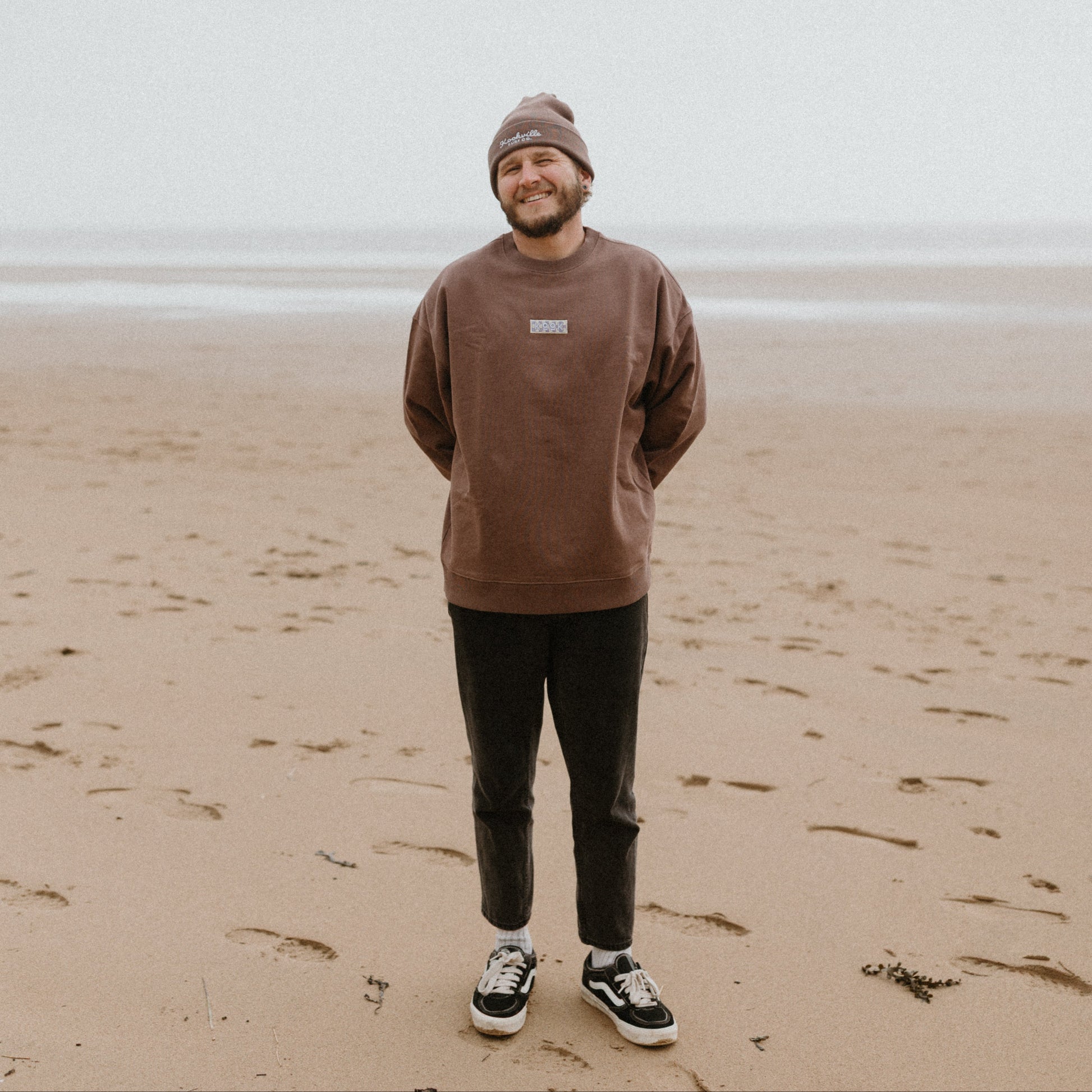 Man standing on a beach wearing a brown sweatshirt and beanie.