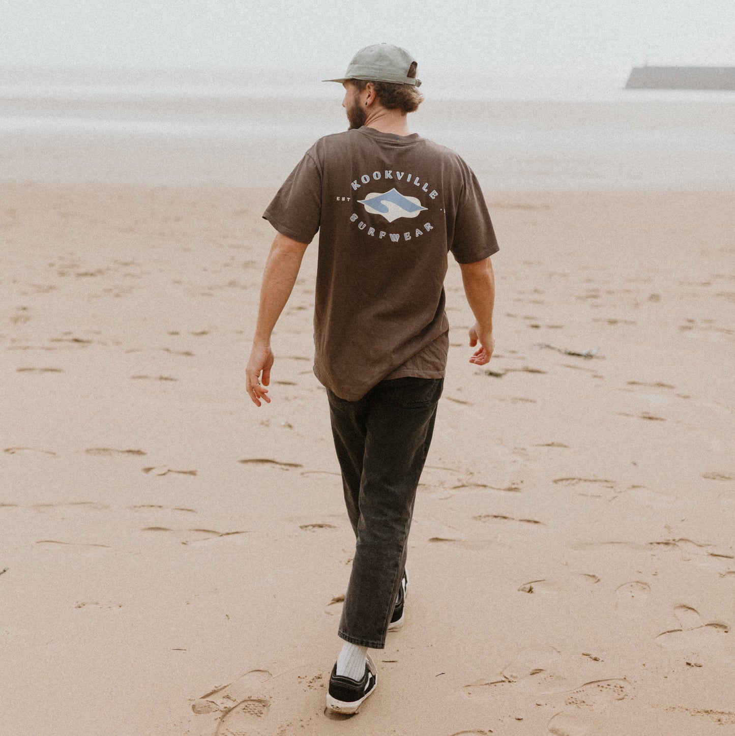 Man walking on a beach wearing a brown t-shirt with a logo and text.