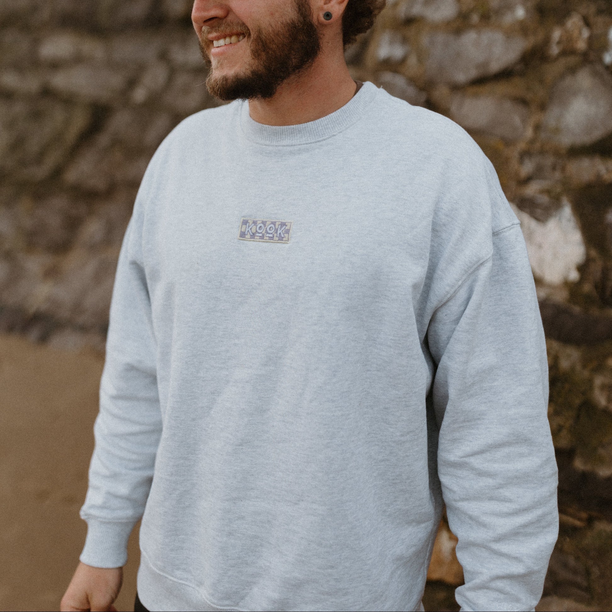 Man wearing a light gray sweatshirt and cap standing against a stone wall.