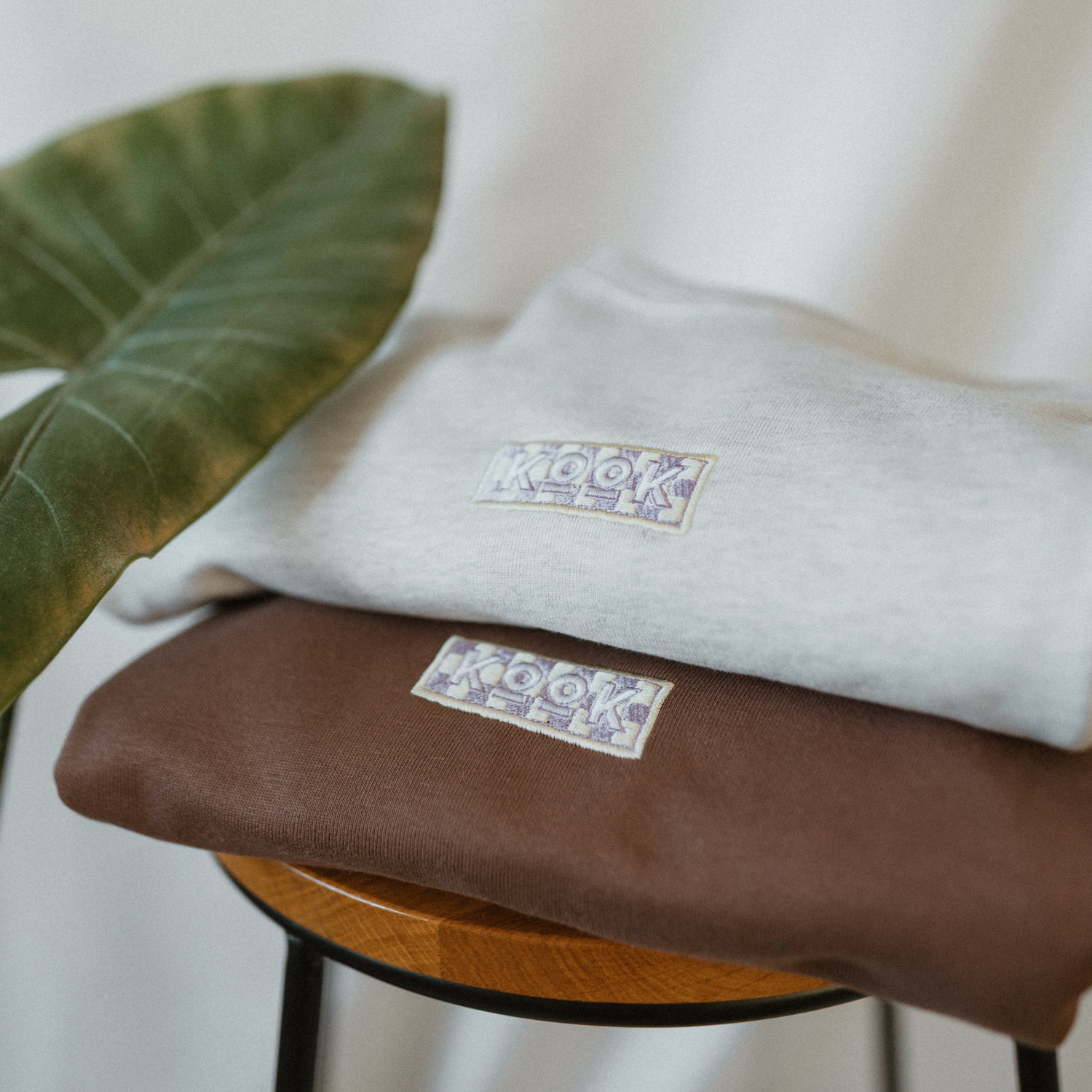 Two folded sweaters, one beige and one brown, with a brand logo on a white background.