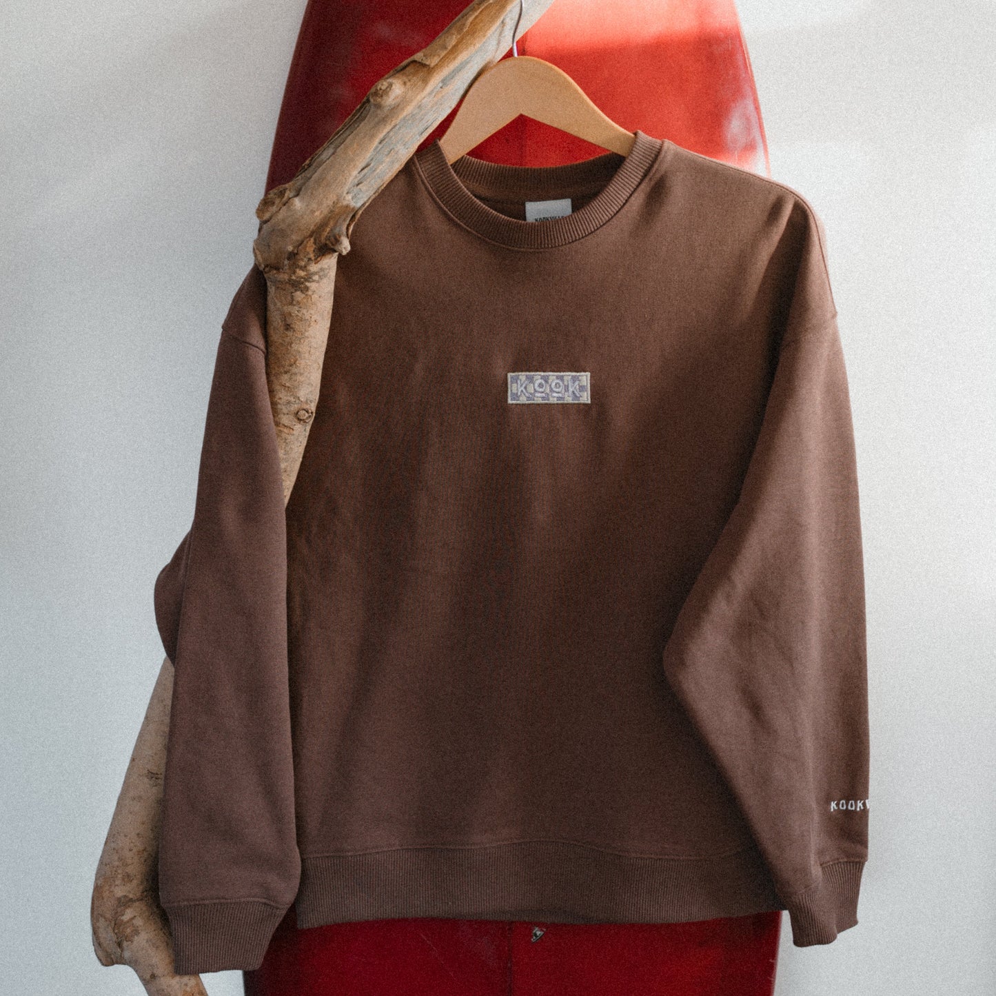 Brown sweatshirt with a small logo on a hanger against a white wall.