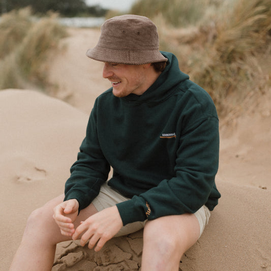Person sitting on a sand dune wearing a green hoodie and brown bucket hat.