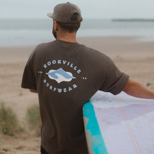 Man holding a surfboard on a beach wearing a 'Kookville Surfwear' t-shirt.
