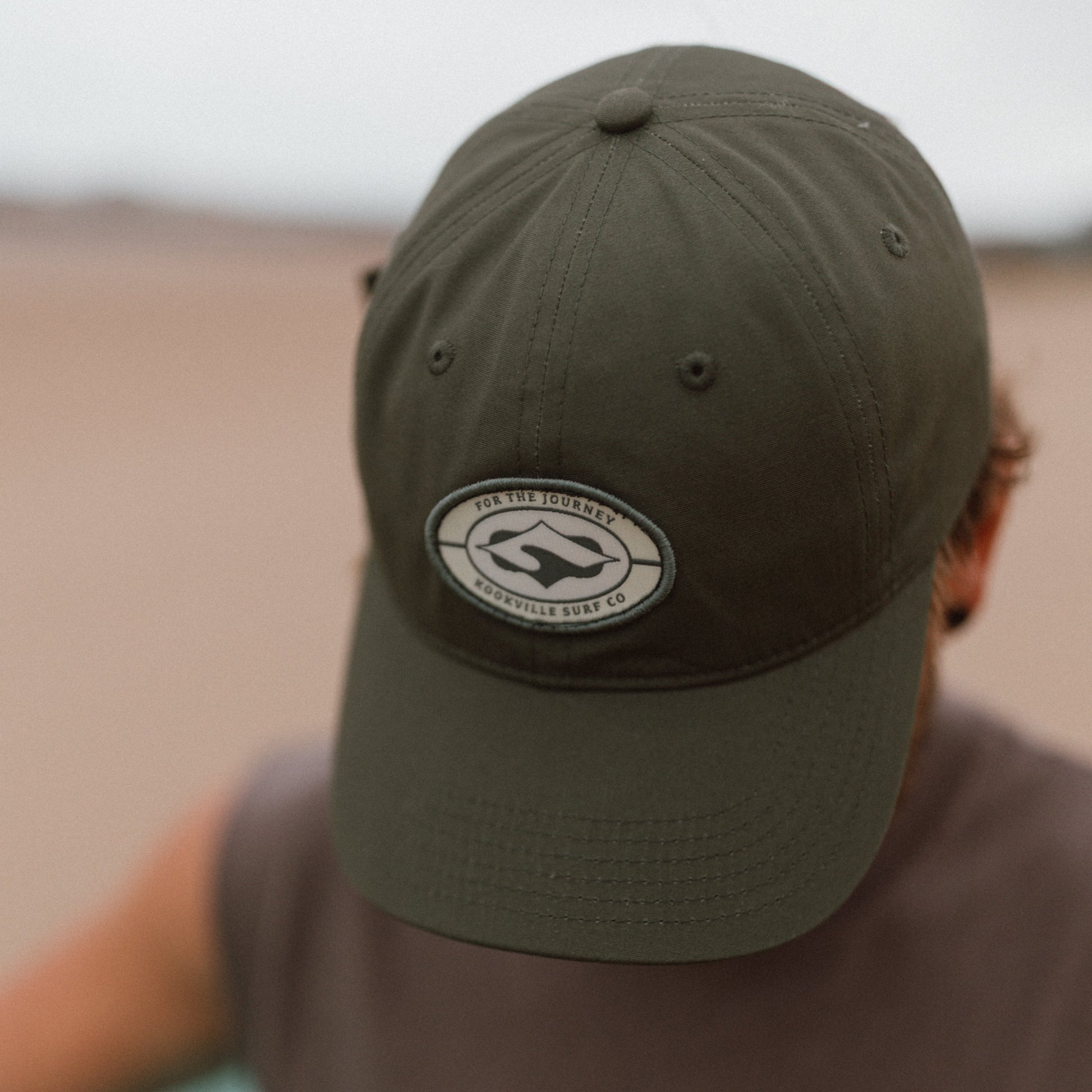 A person wearing an khaki green baseball cap with a circular 'For The Journey' & 'Kookville Surf Co' logo patch on the front, against a blurred beach background.