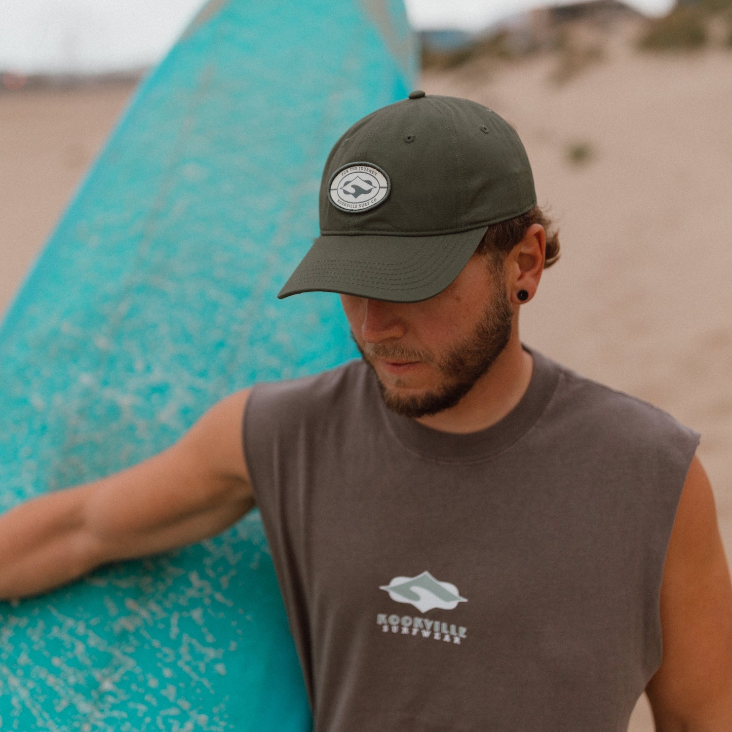 A man on a sandy beach carrying a turquoise surfboard, wearing a dark olive green baseball cap with a circular Kookville logo and a sleeveless grey t-shirt with a white graphic logo on the chest.
